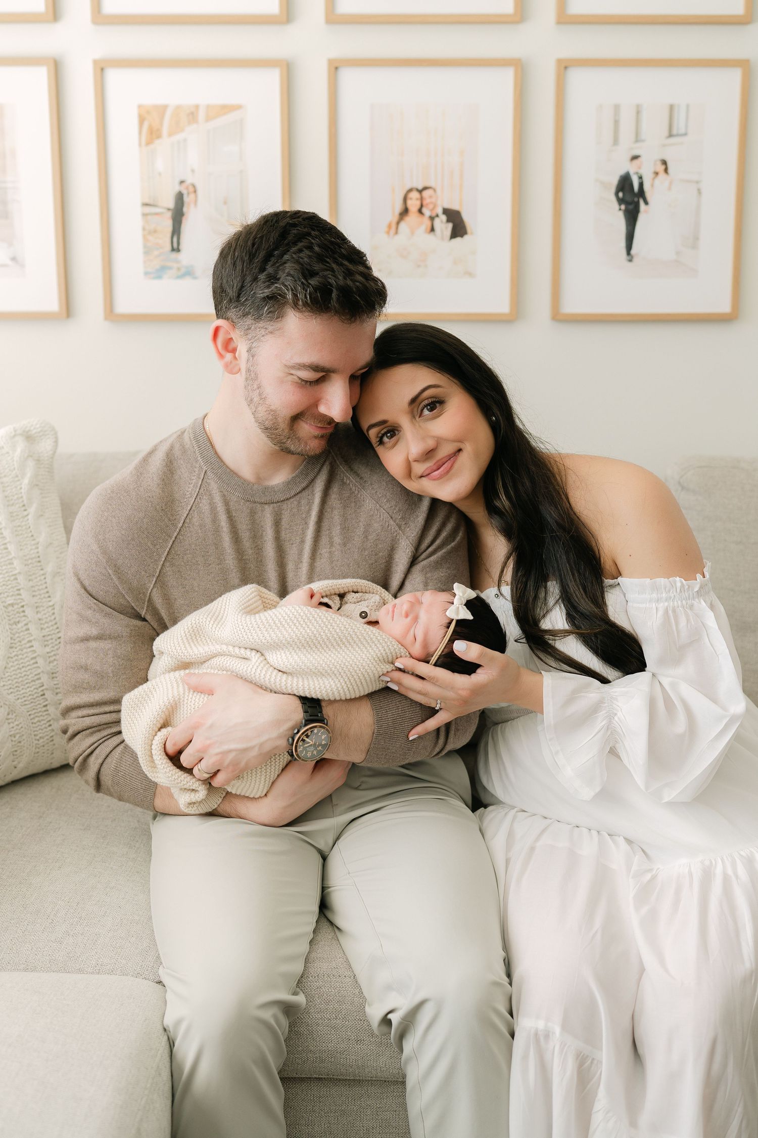 A couple sits on a beige sofa together, smiling down at their newborn baby wrapped in a cream blanket.
