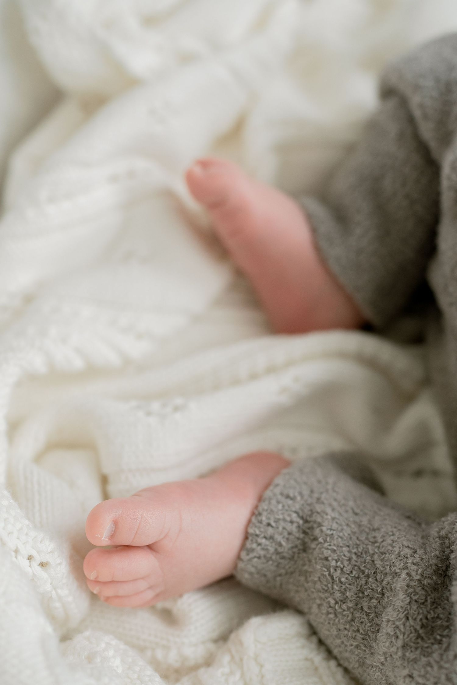 Close-up of tiny newborn baby feet resting on a soft white knit blanket with gray pants.