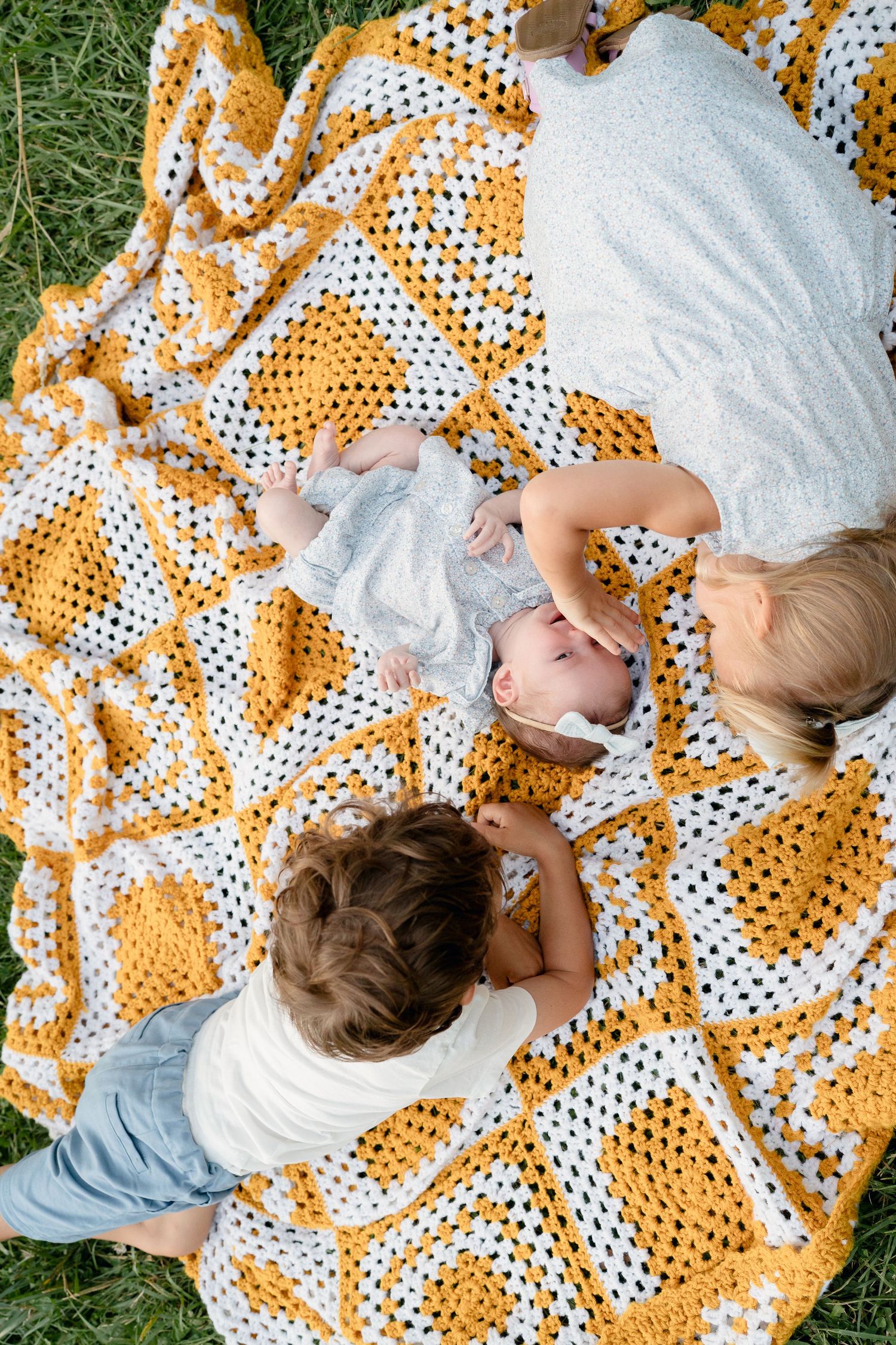 Mother and children relaxing on a yellow and white crocheted blanket on green grass outdoors.