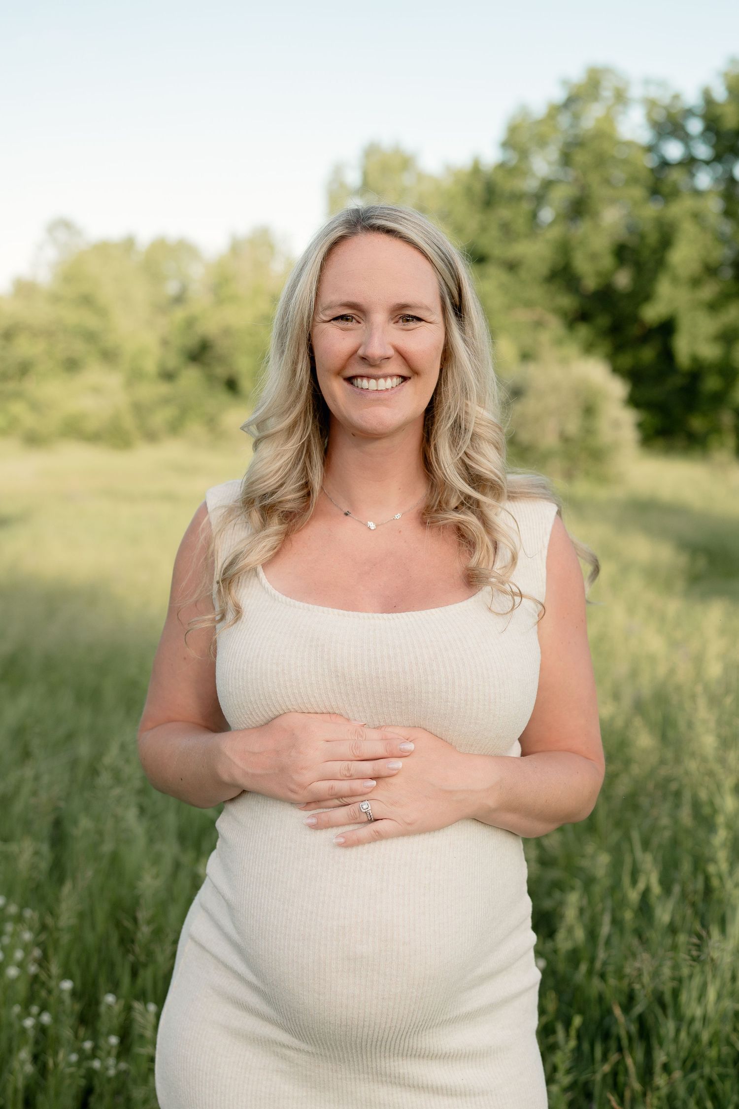 Smiling pregnant woman in cream ribbed dress cradling baby bump outdoors in lush green field during golden hour.