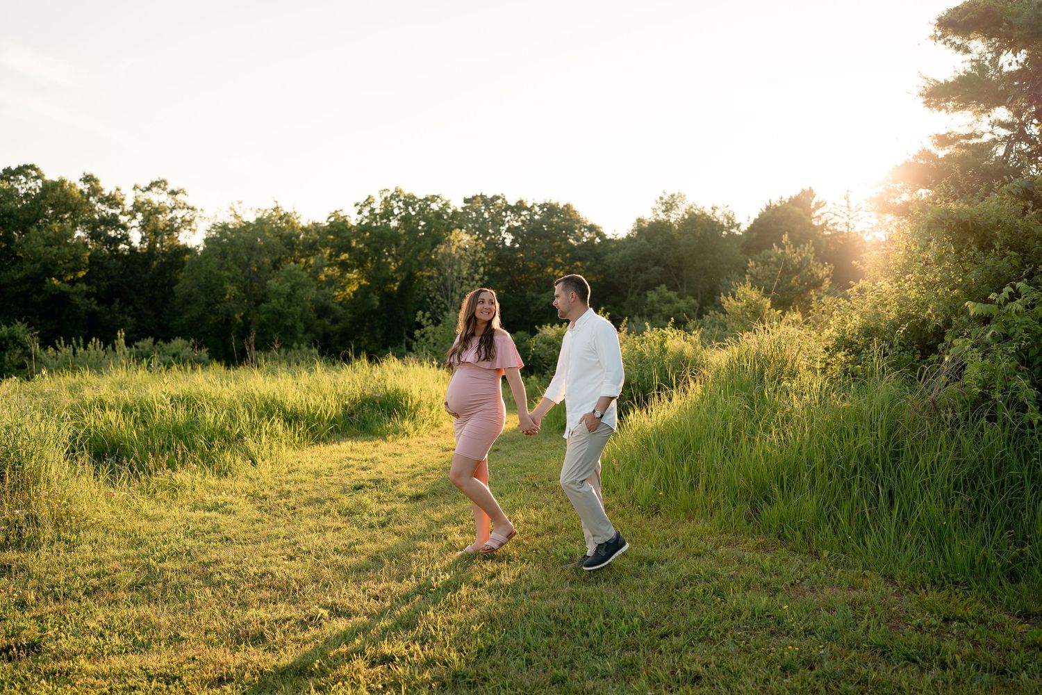 Happy pregnant couple playfully dancing in a sunny green meadow at sunset.