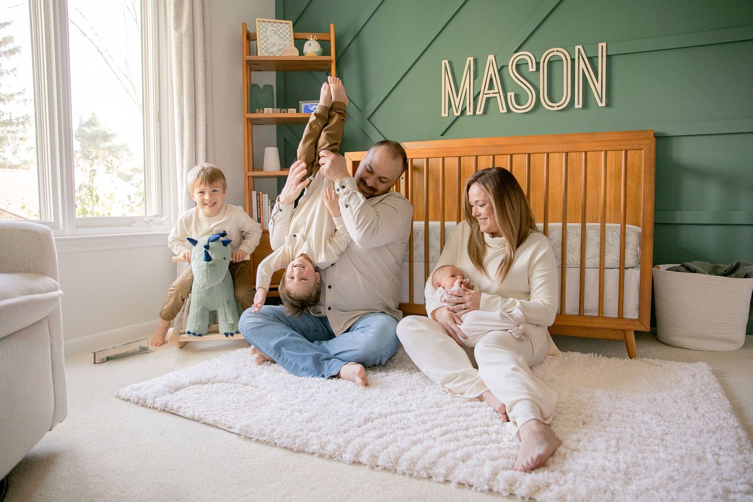 Family of four sits on nursery floor beneath name sign MASON, with dad holding toddler and mom cradling newborn.