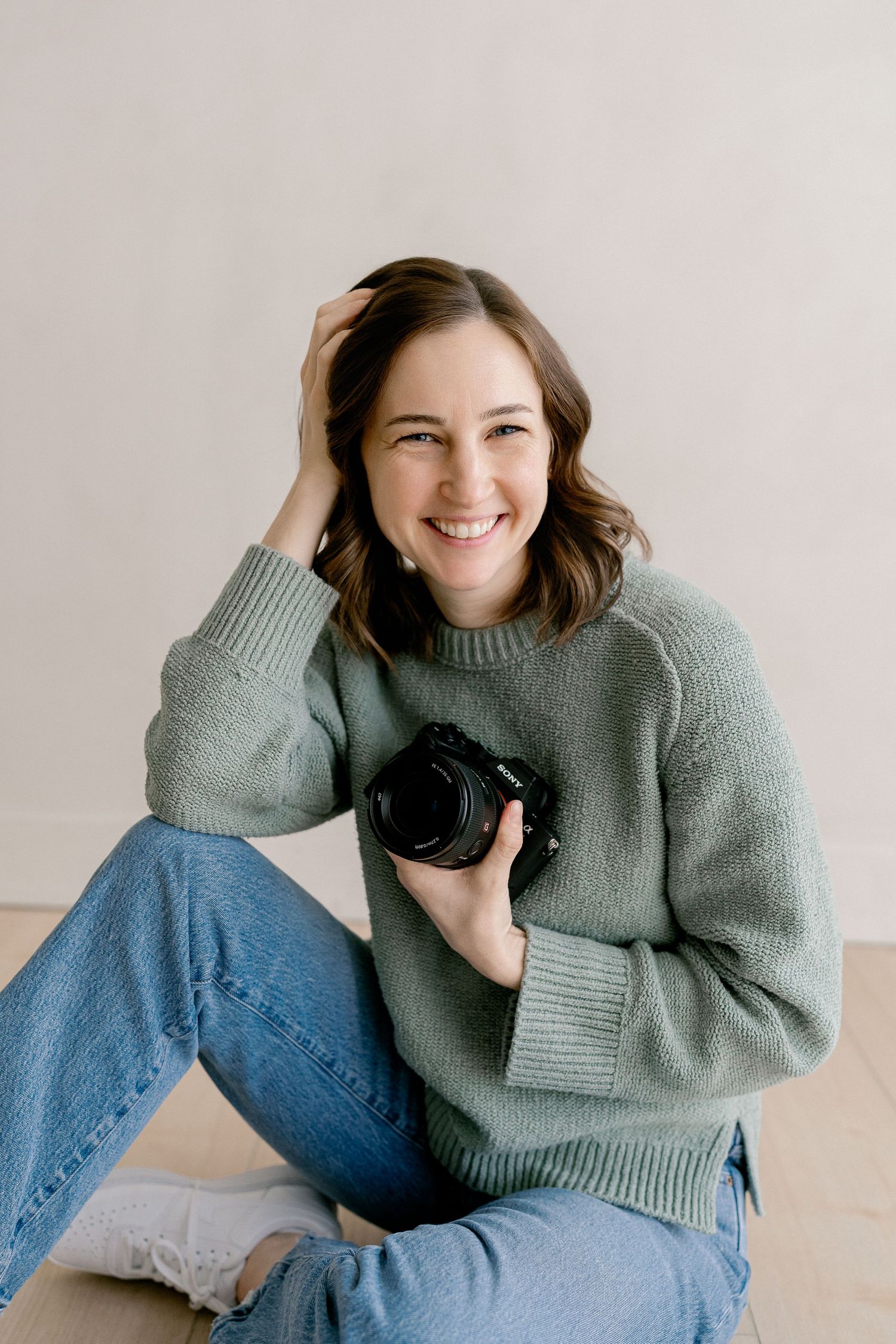 Smiling female photographer sits casually holding a camera, wearing a green sweater and jeans indoors.