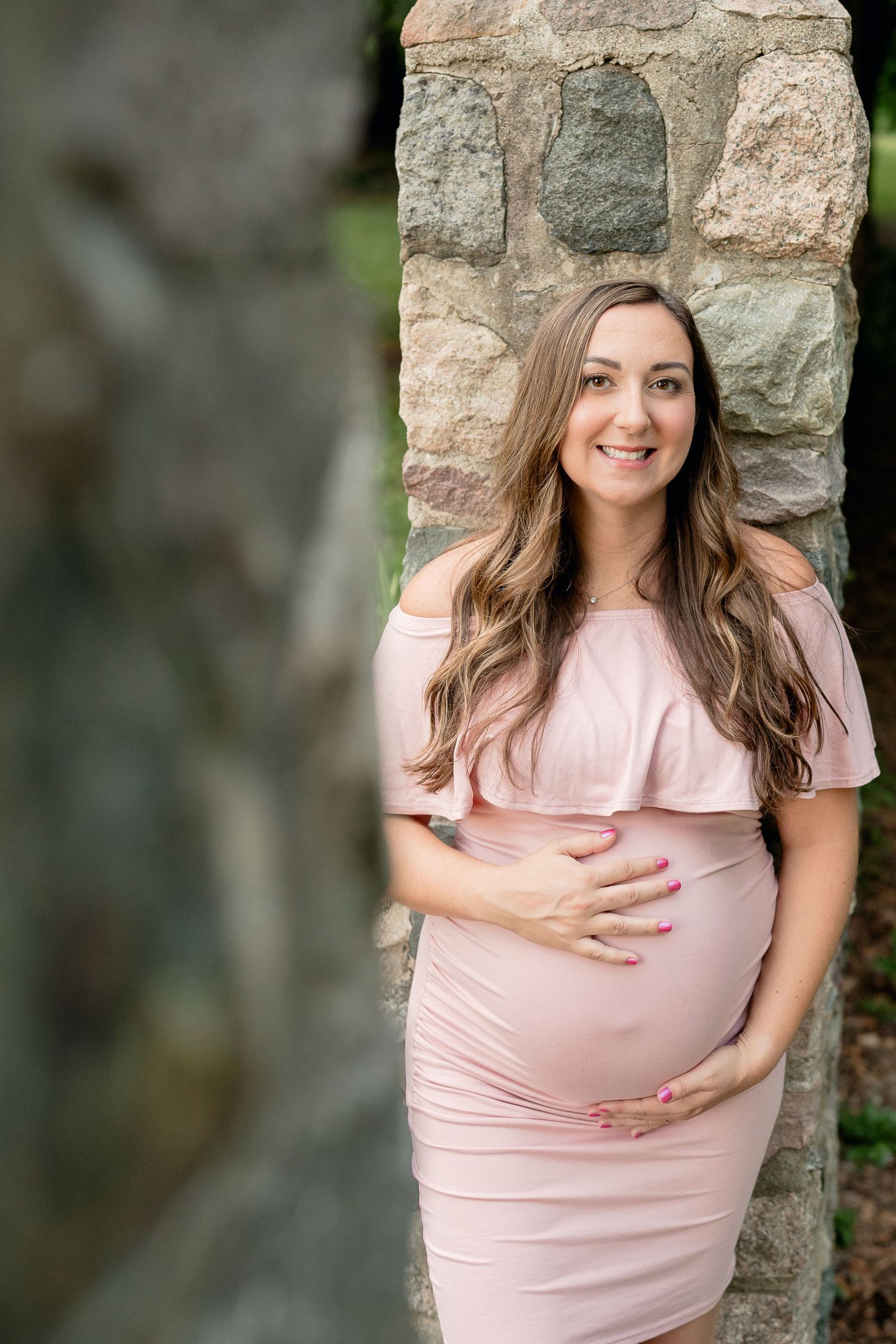 Smiling pregnant woman in a blush off-shoulder dress leaning against a stone wall during outdoor maternity session.