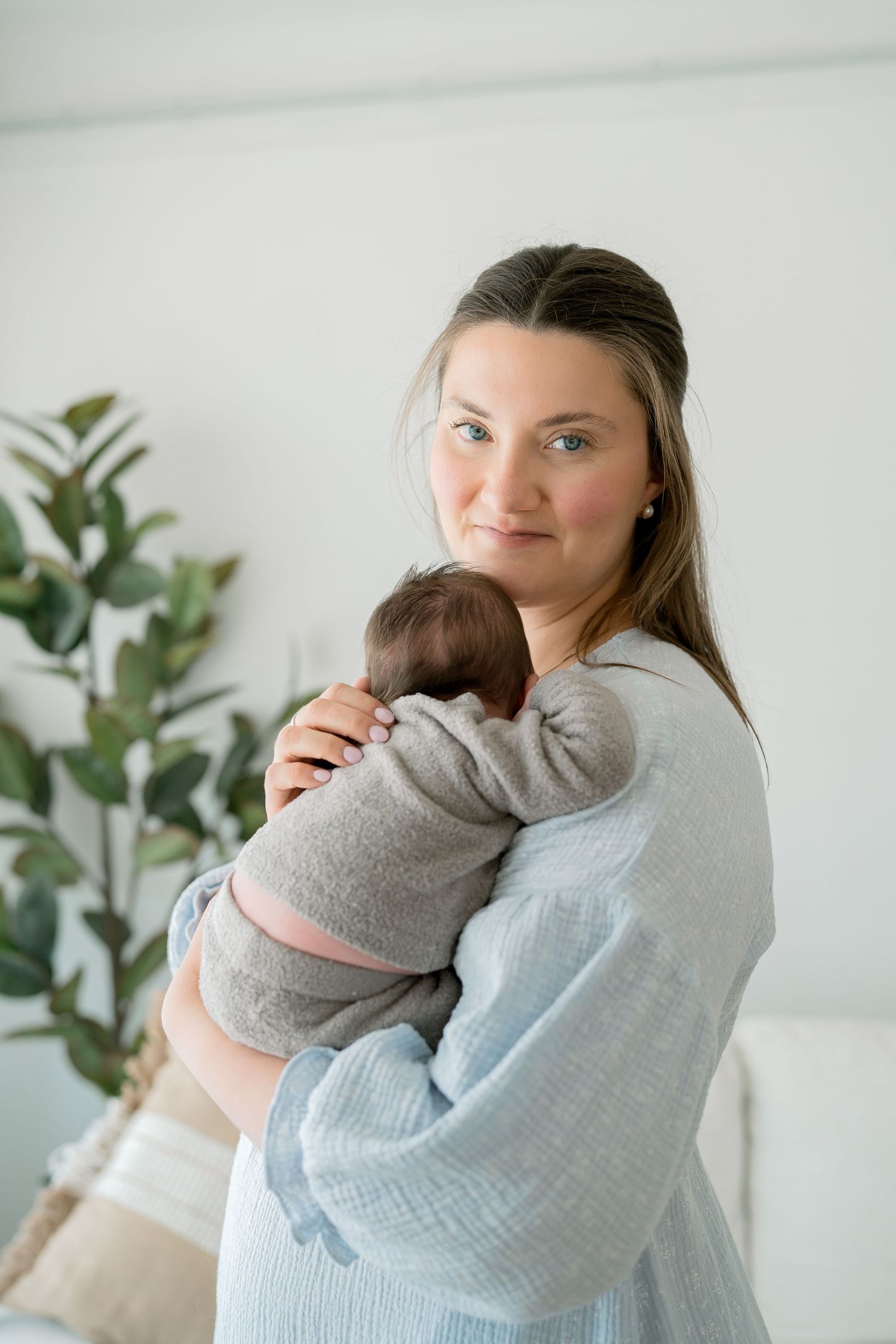 A mother tenderly holds her newborn baby while looking at the camera in a bright, minimalist white room.