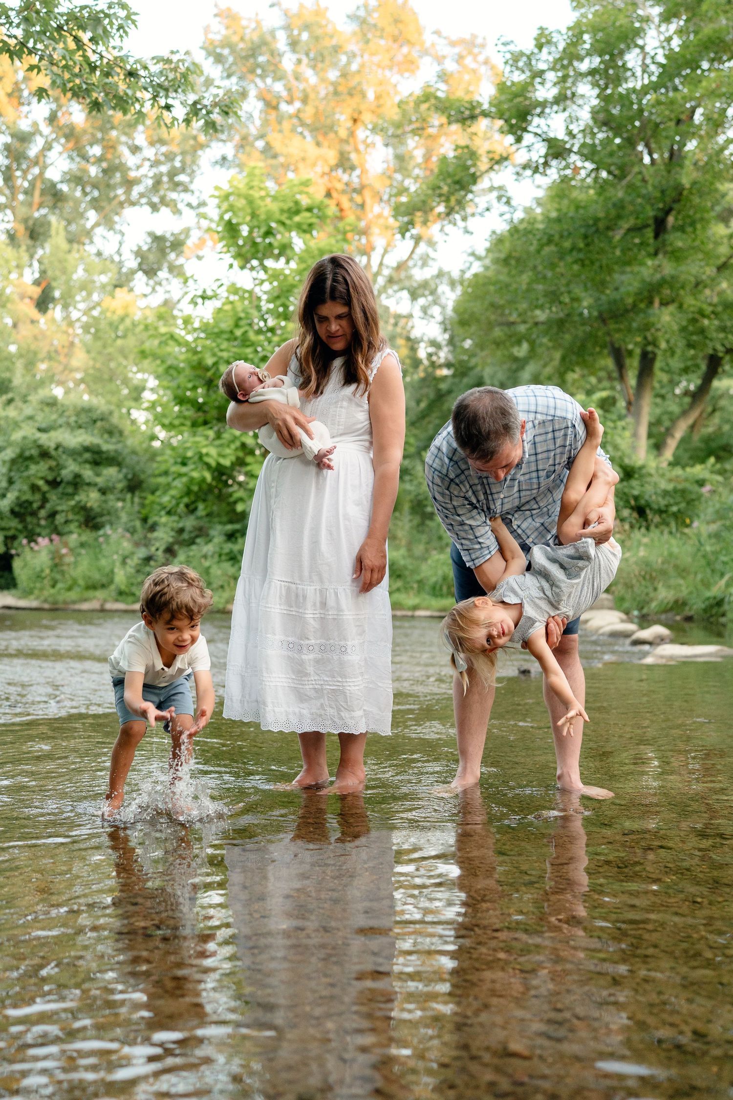 Family of five enjoying a sunny day wading in a shallow creek surrounded by lush green trees.