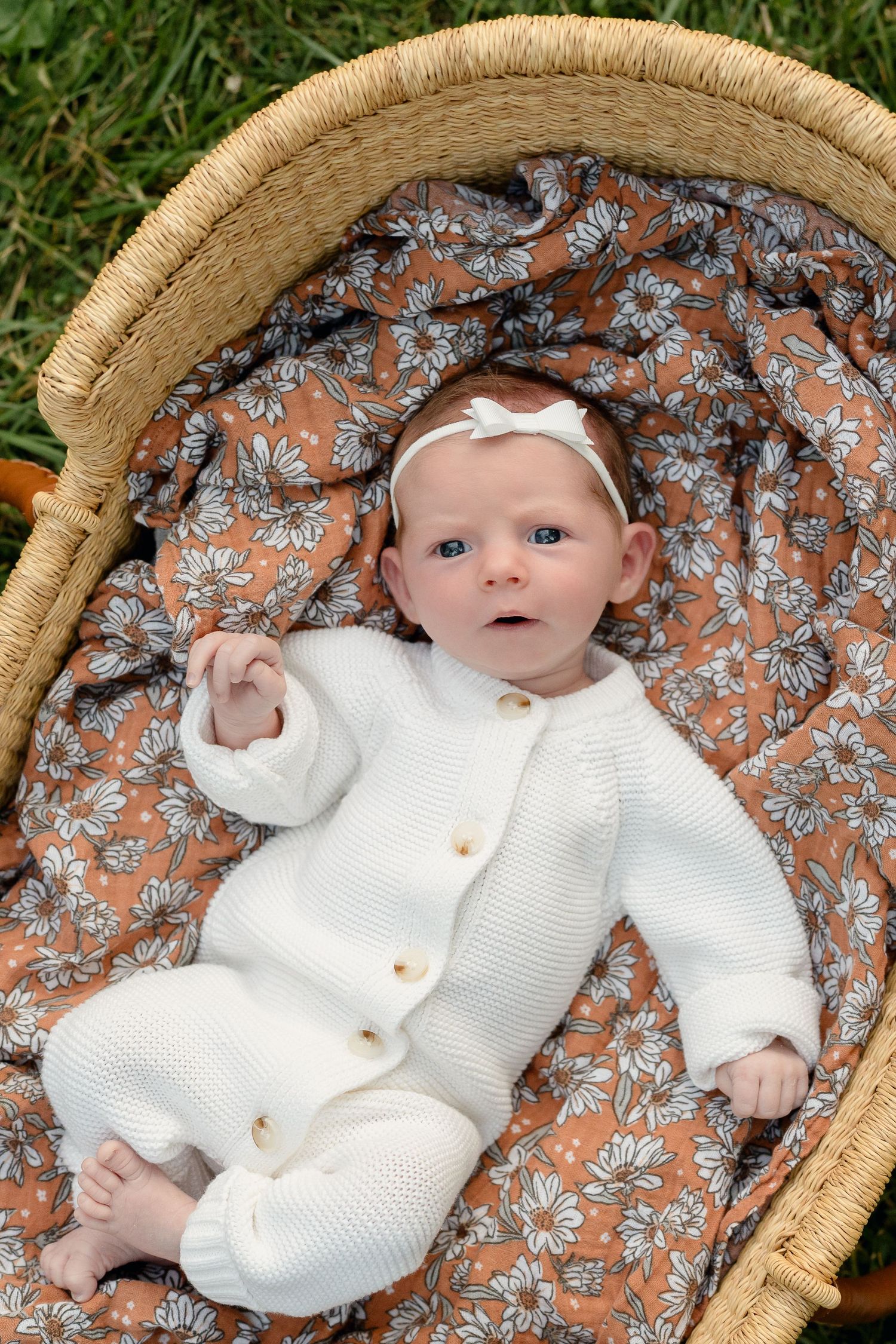 Newborn baby in white knit romper lying in a wicker Moses basket with floral lining outdoors.