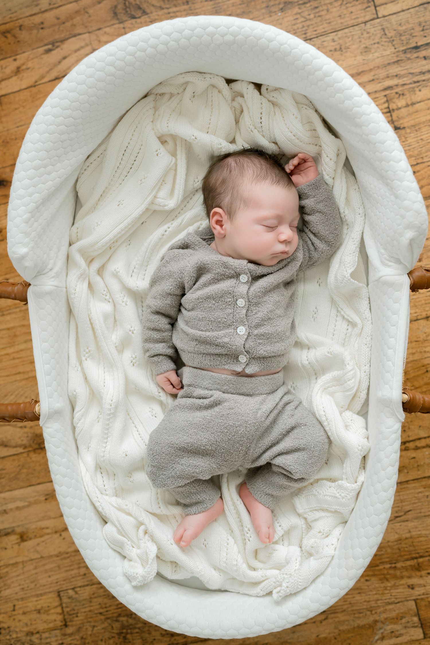 Sleeping newborn baby in gray knit outfit lying in white oval bassinet on cream blanket.