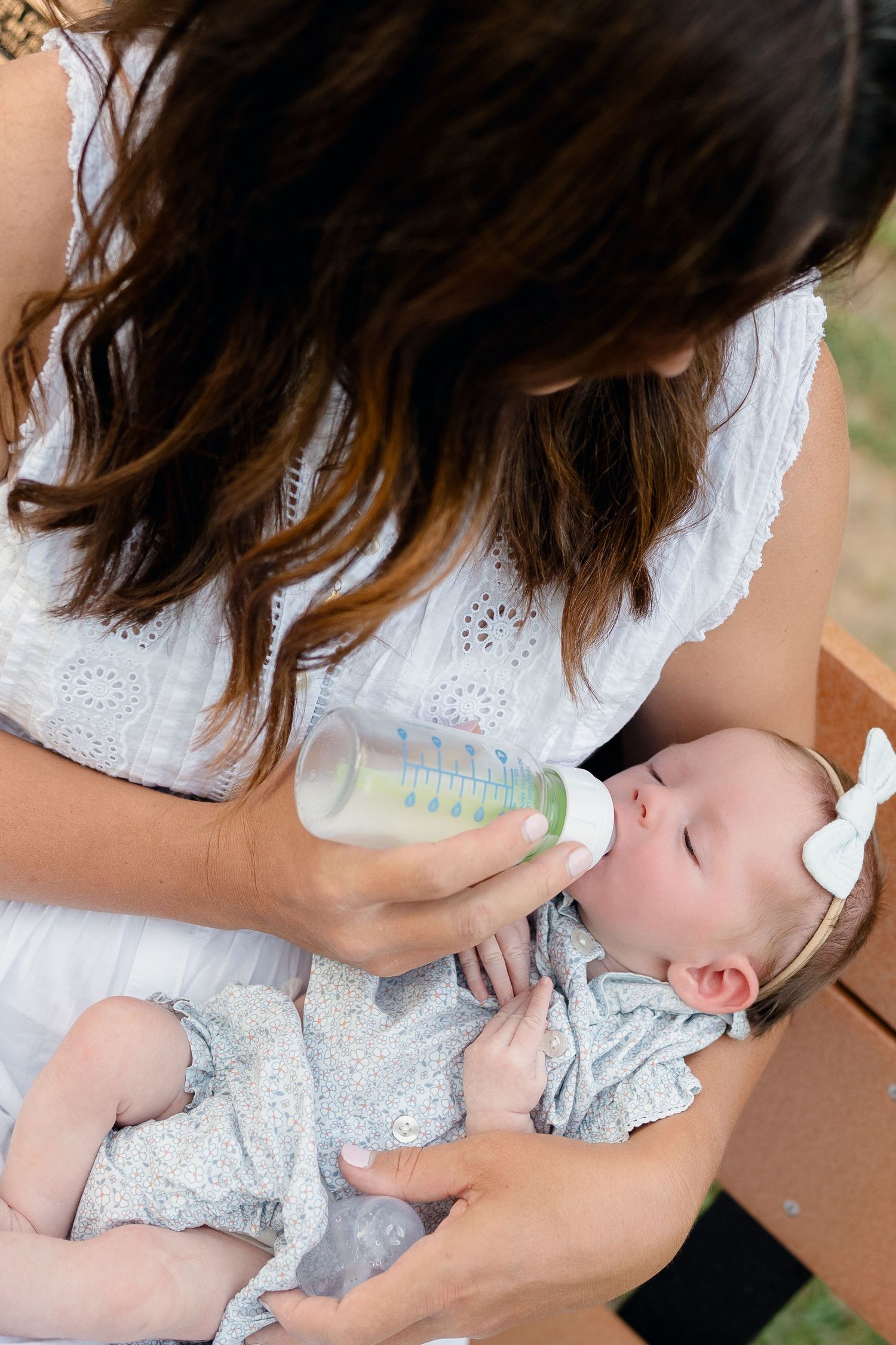 A mother in a white dress bottle-feeding her infant baby outdoors, with the baby wearing a gray outfit and white bow headband.