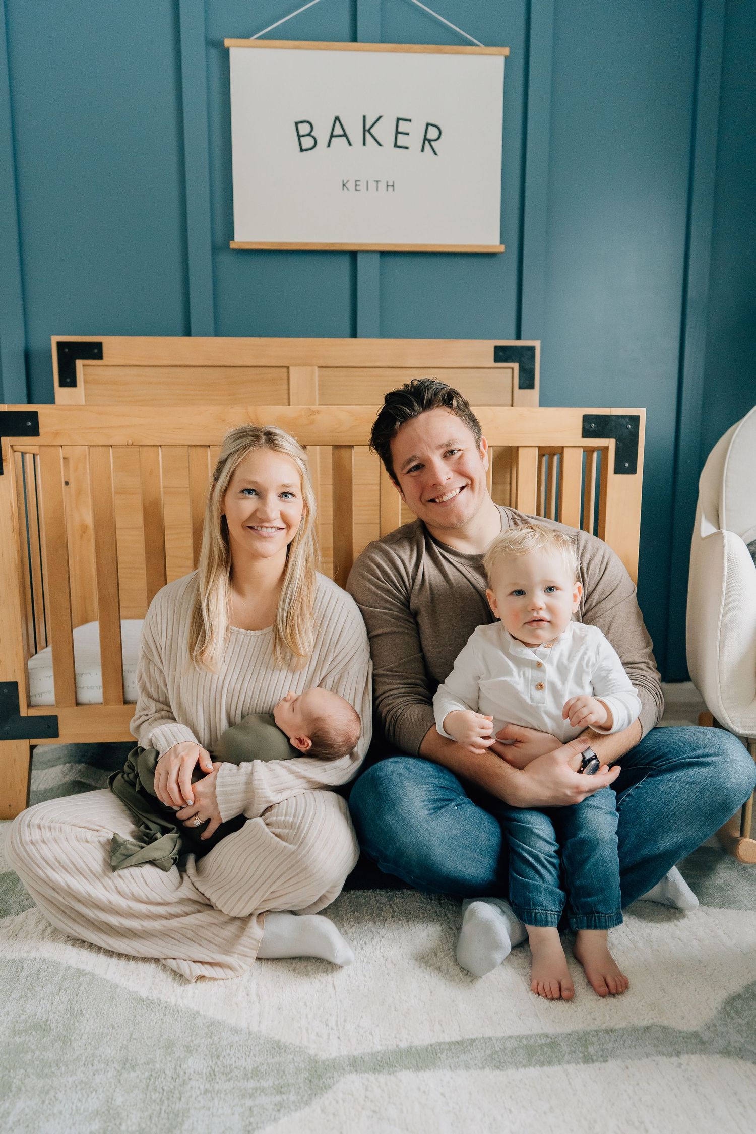 Happy family with newborn and toddler sitting in front of wooden crib in nursery.