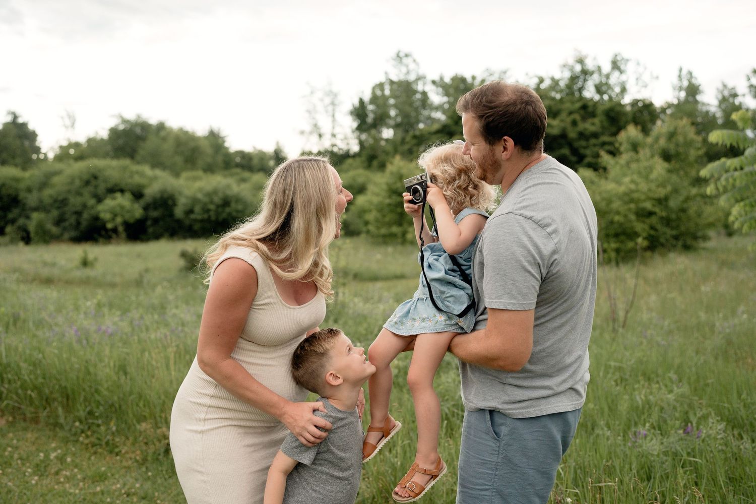 Family of four posing outdoors in green field, parents with young boy and girl holding camera, candid lifestyle portrait.