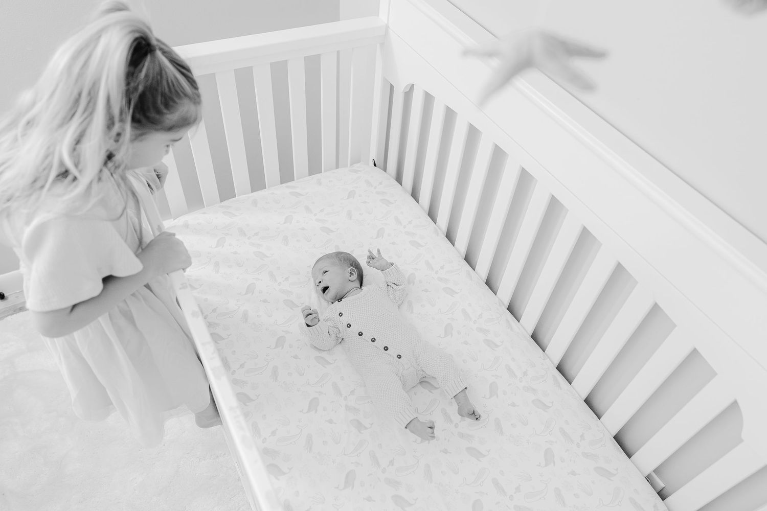 Black and white photo of a young girl peering into a white crib at her newborn baby sibling.