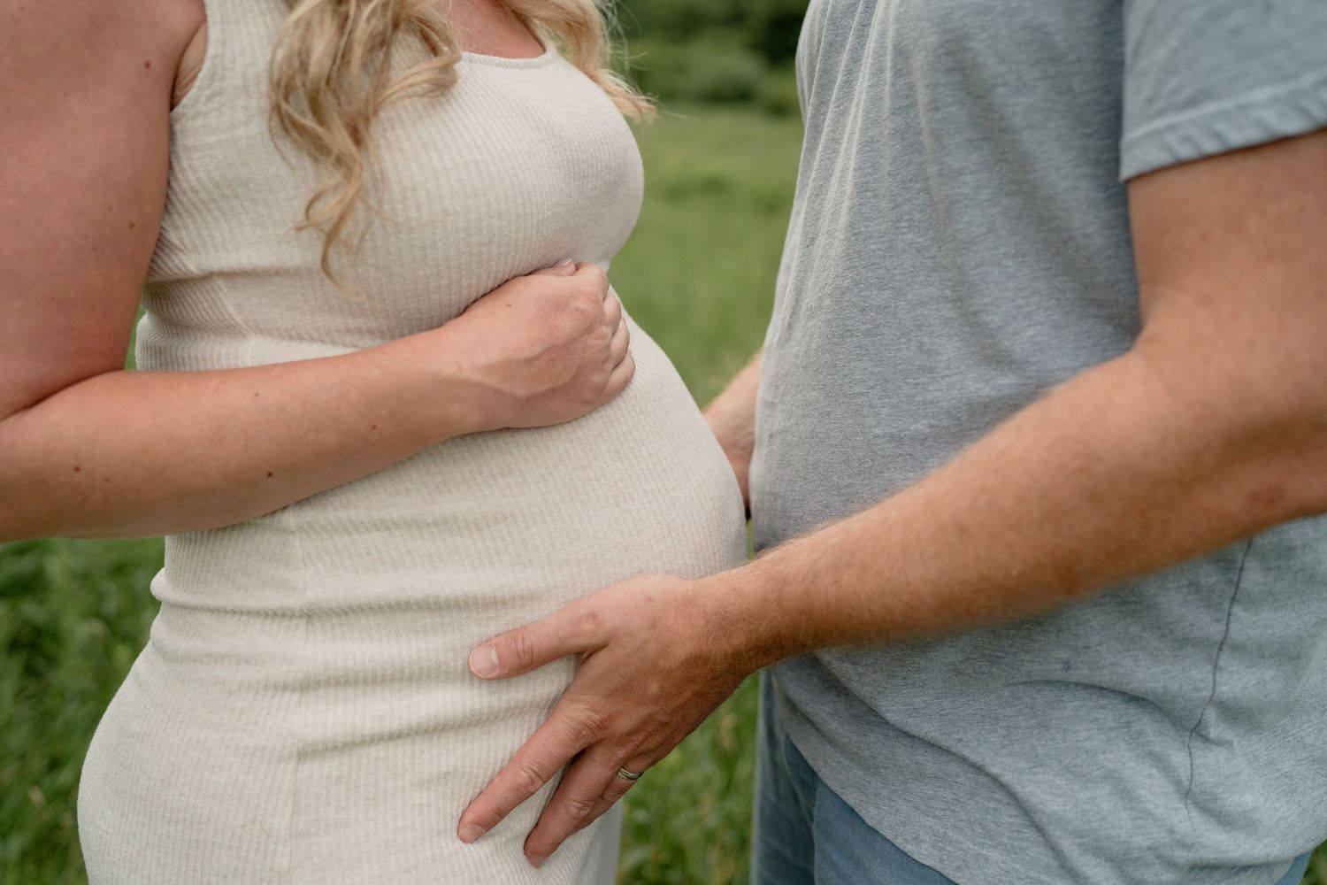 Pregnant woman in cream dress with partner's hand on baby bump outdoors in nature setting.