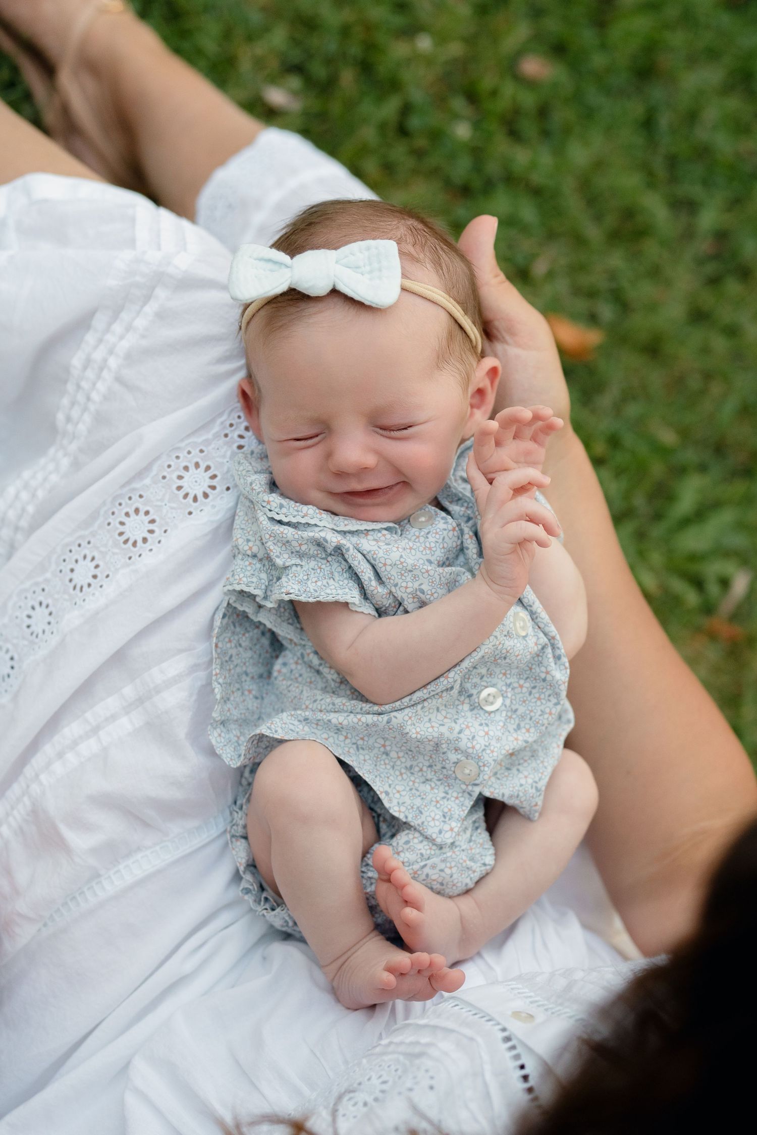 Smiling newborn baby girl in floral romper and white bow headband resting on mother's lap outdoors.