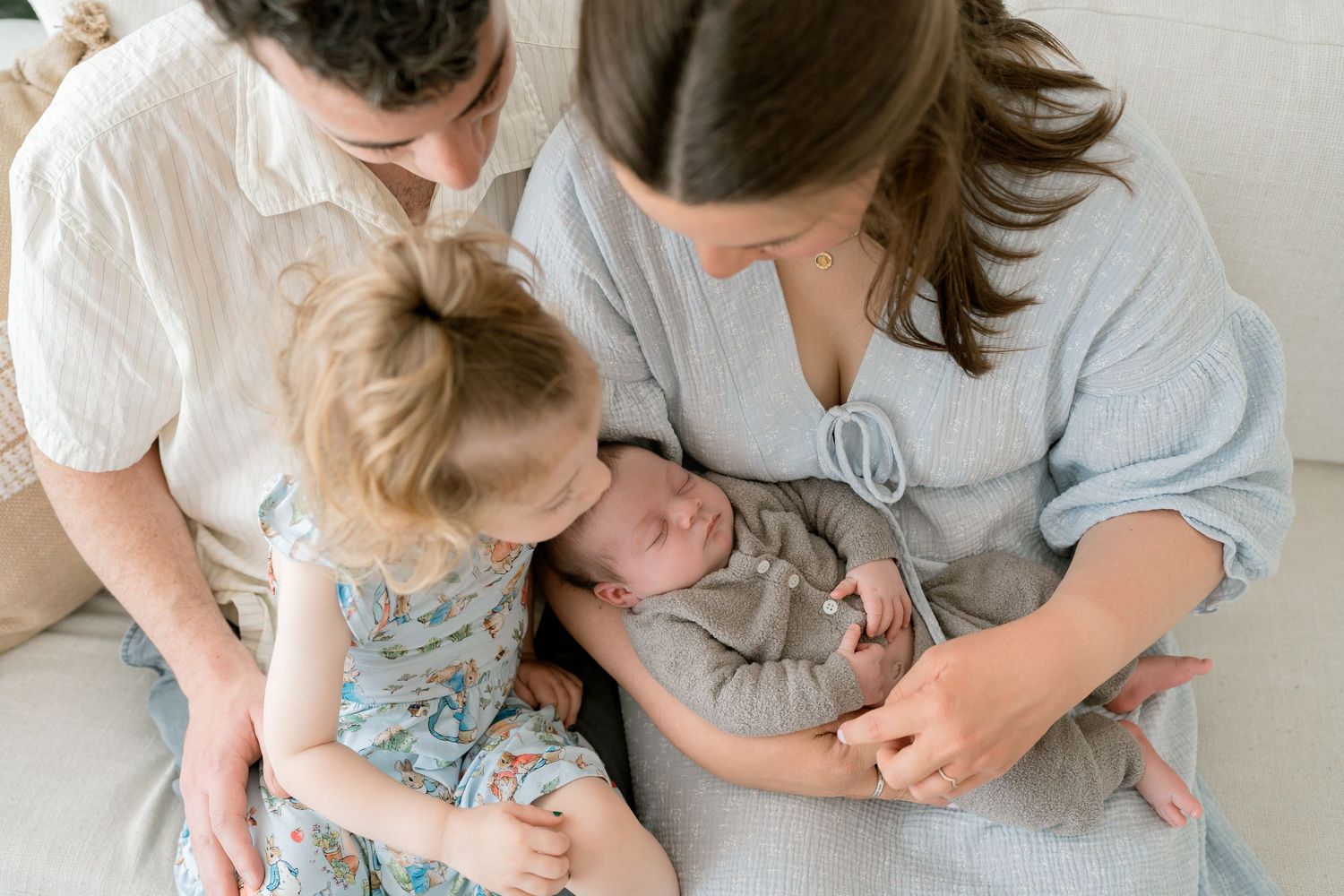 Two mothers and a toddler girl lovingly gaze at a sleeping newborn baby during a family portrait session.