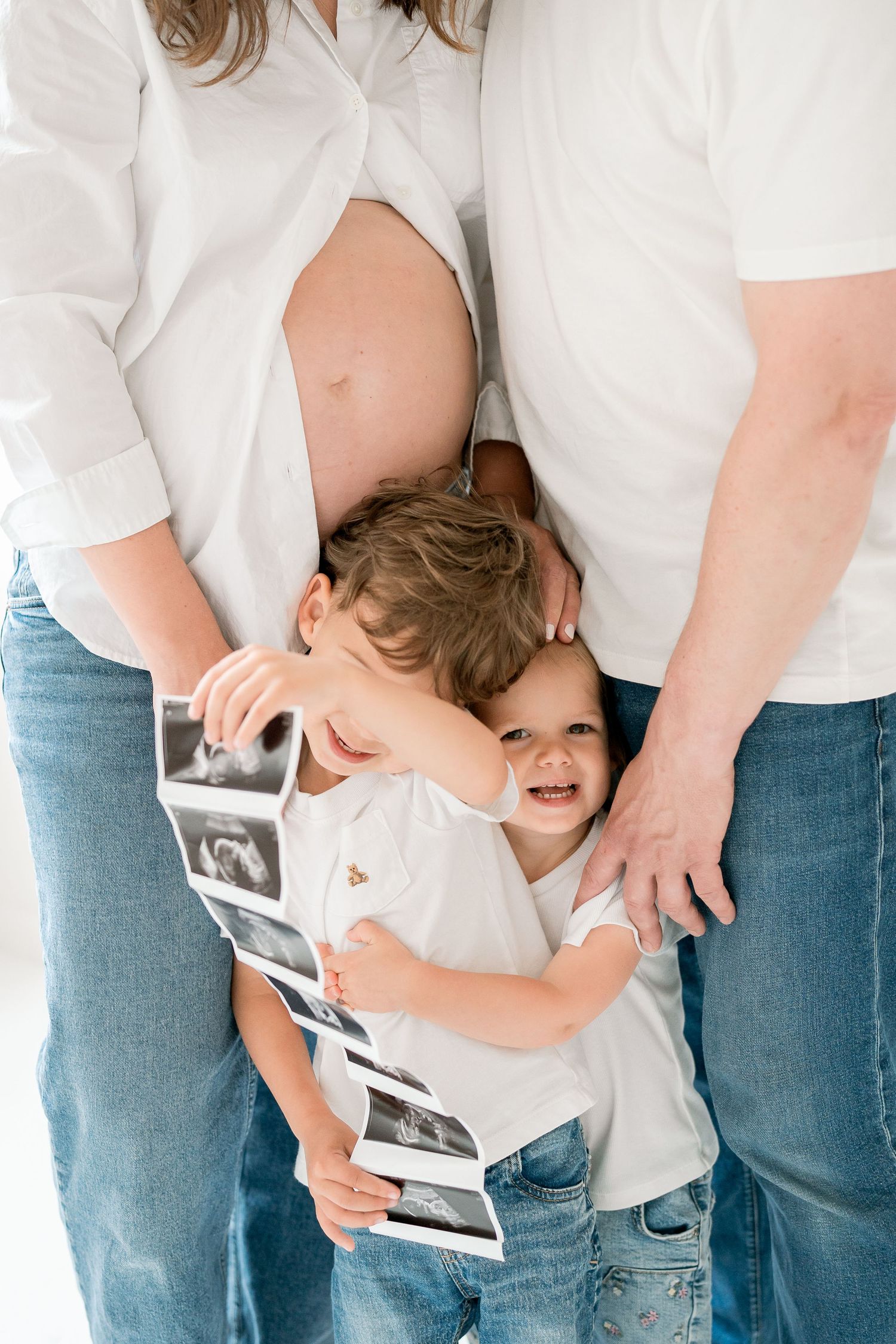 Two young children hugging pregnant mother's belly while holding ultrasound photos during maternity photo session.