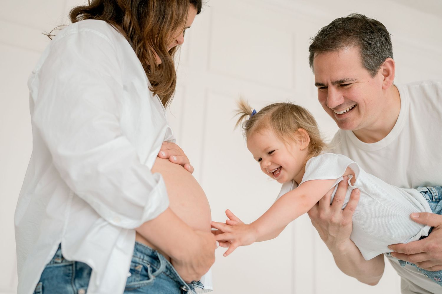 Smiling father holding toddler daughter who touches pregnant mother's bare belly in a bright white room.