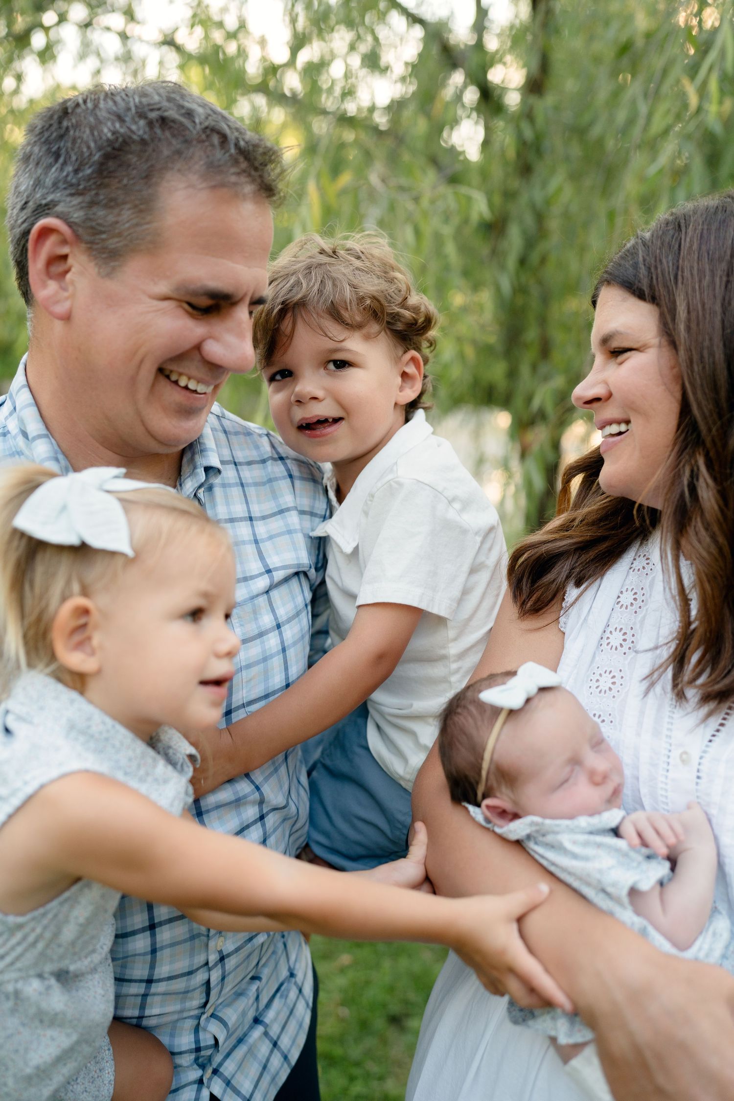 Happy family with toddler girl, young boy, and newborn baby laughing together outdoors near willow tree.