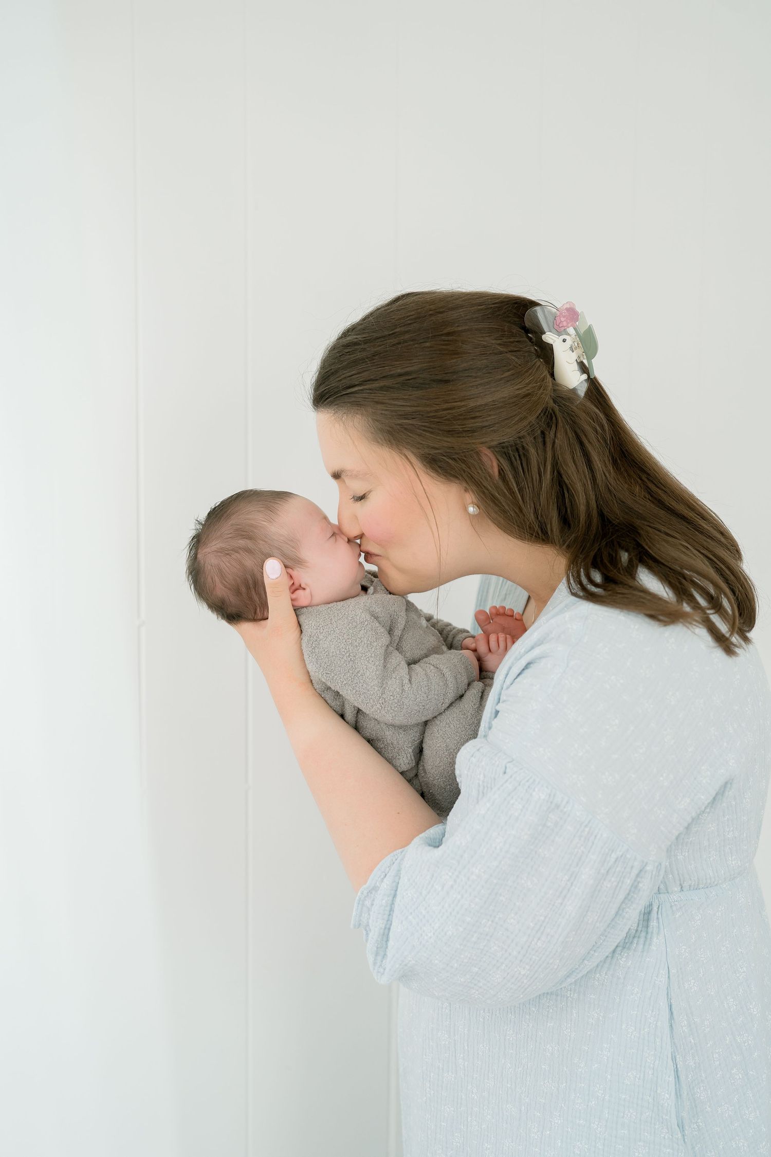 A young mother with a hair clip gently kisses her newborn baby dressed in gray, against a white background.