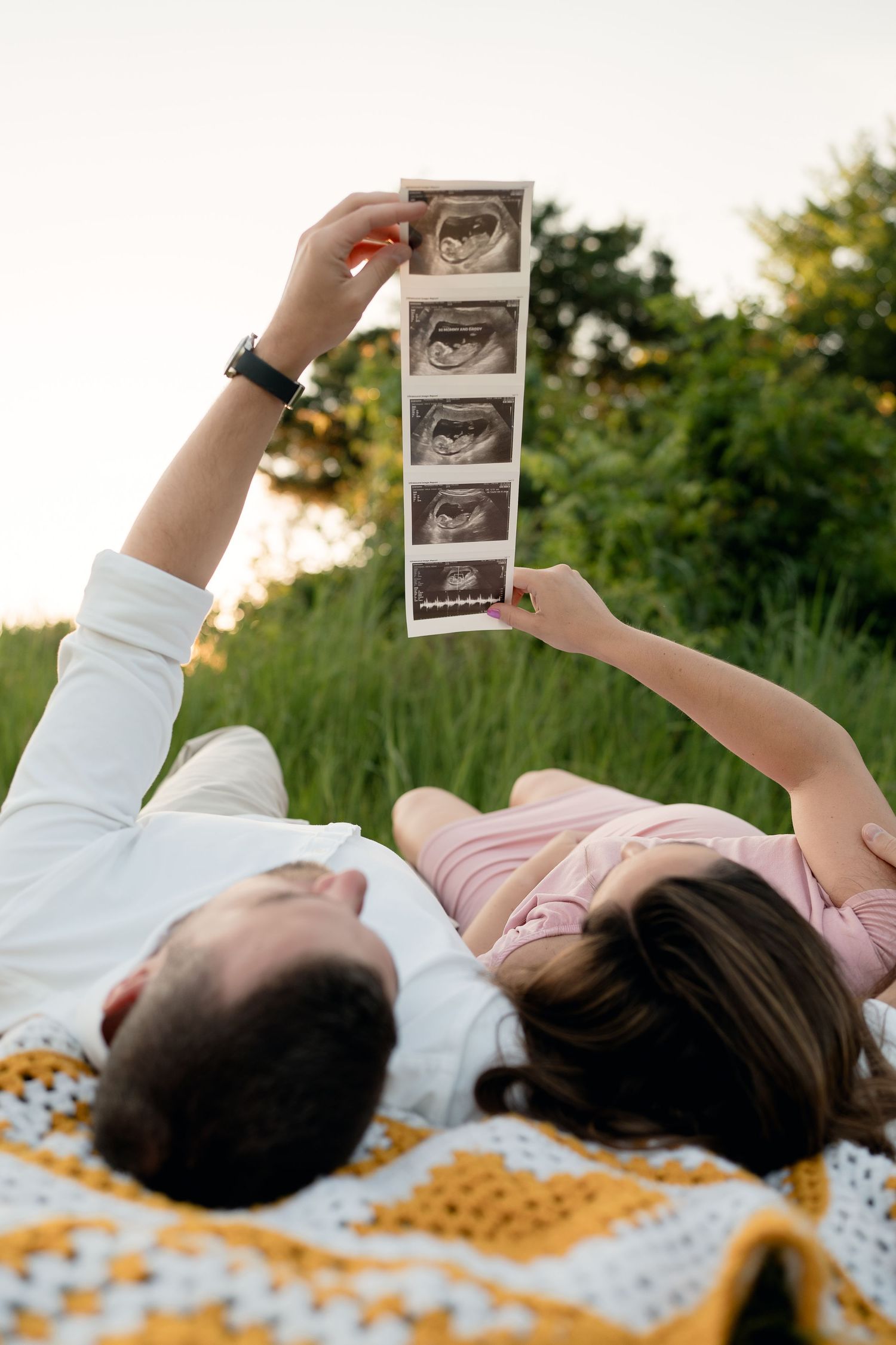 Couple lying in grass holding up ultrasound photos together outdoors.