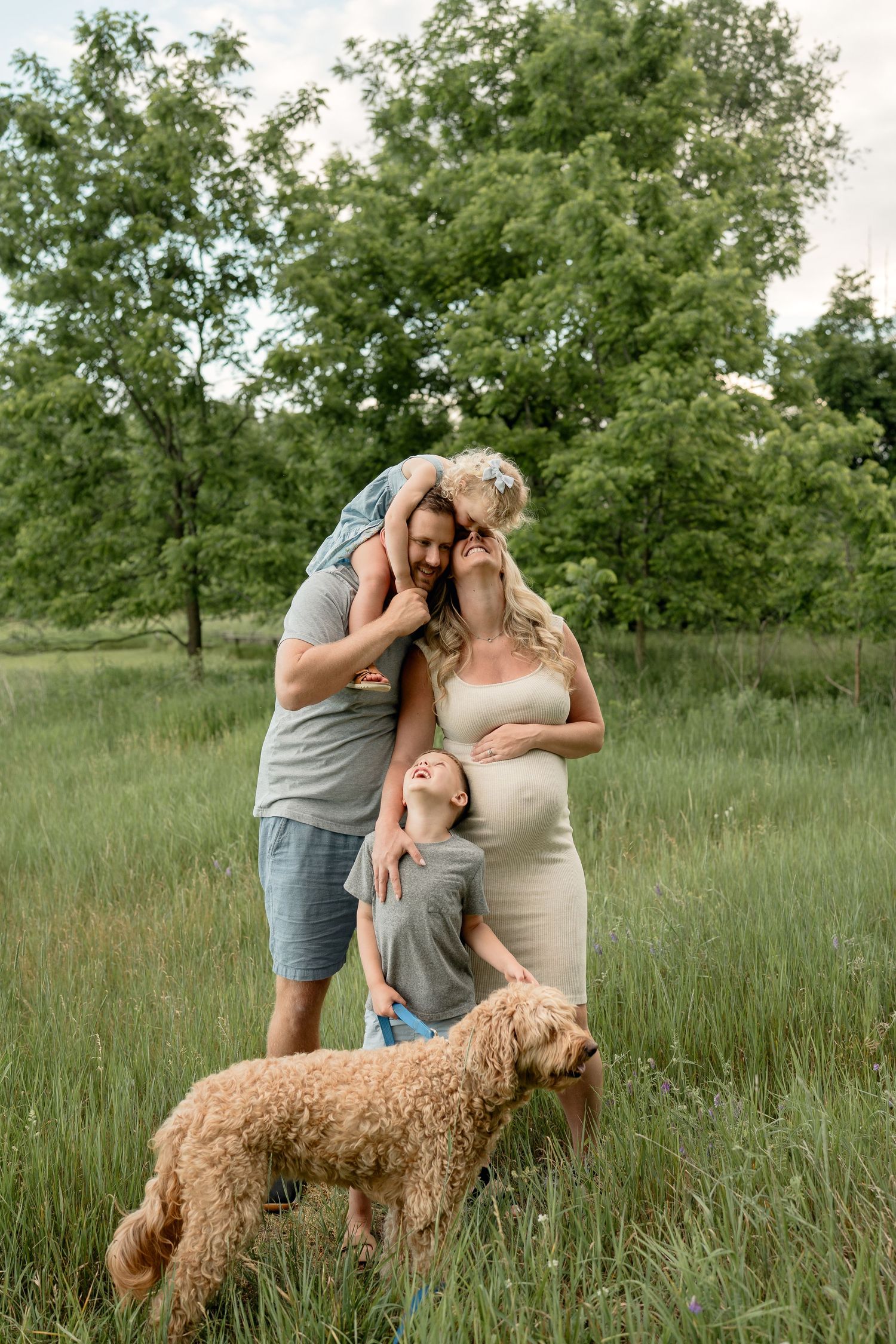 A happy pregnant family with two kids and a golden doodle dog poses together in a lush green meadow.