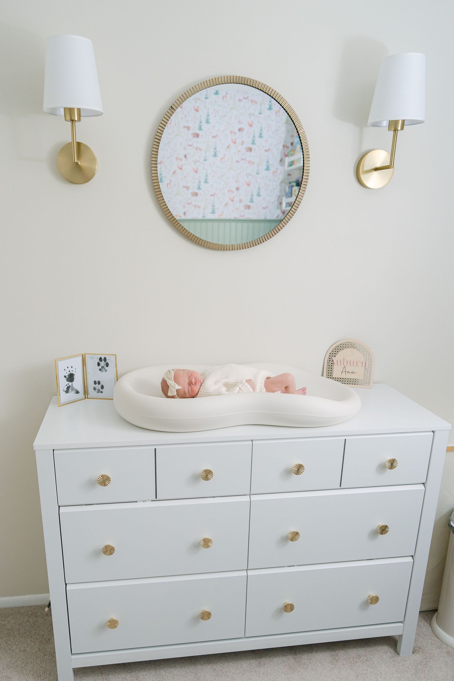 Newborn baby resting on a changing pad atop a white dresser in a softly decorated nursery with gold accents.