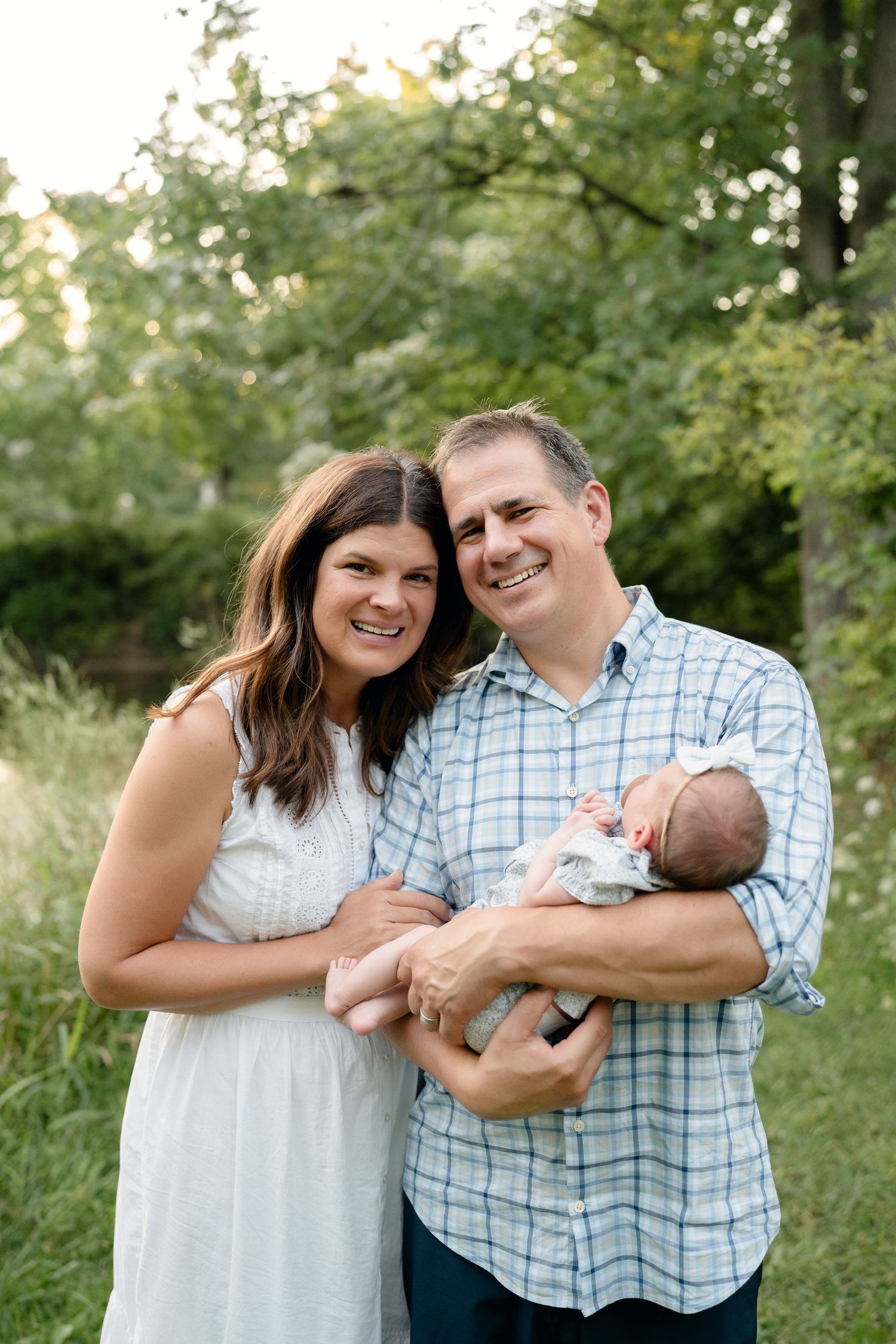Smiling couple holding newborn baby outdoors in lush green park setting, family portrait photography.