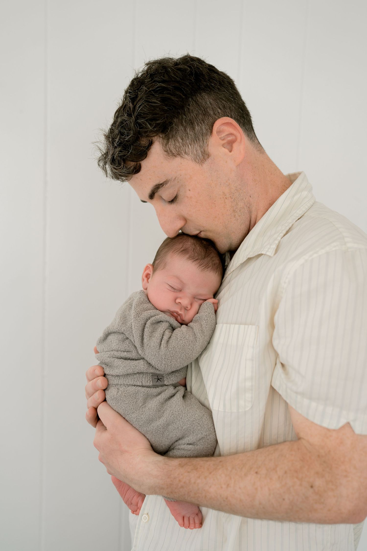 A father tenderly kisses his sleeping newborn baby dressed in gray, cradled gently in his arms against a white background.