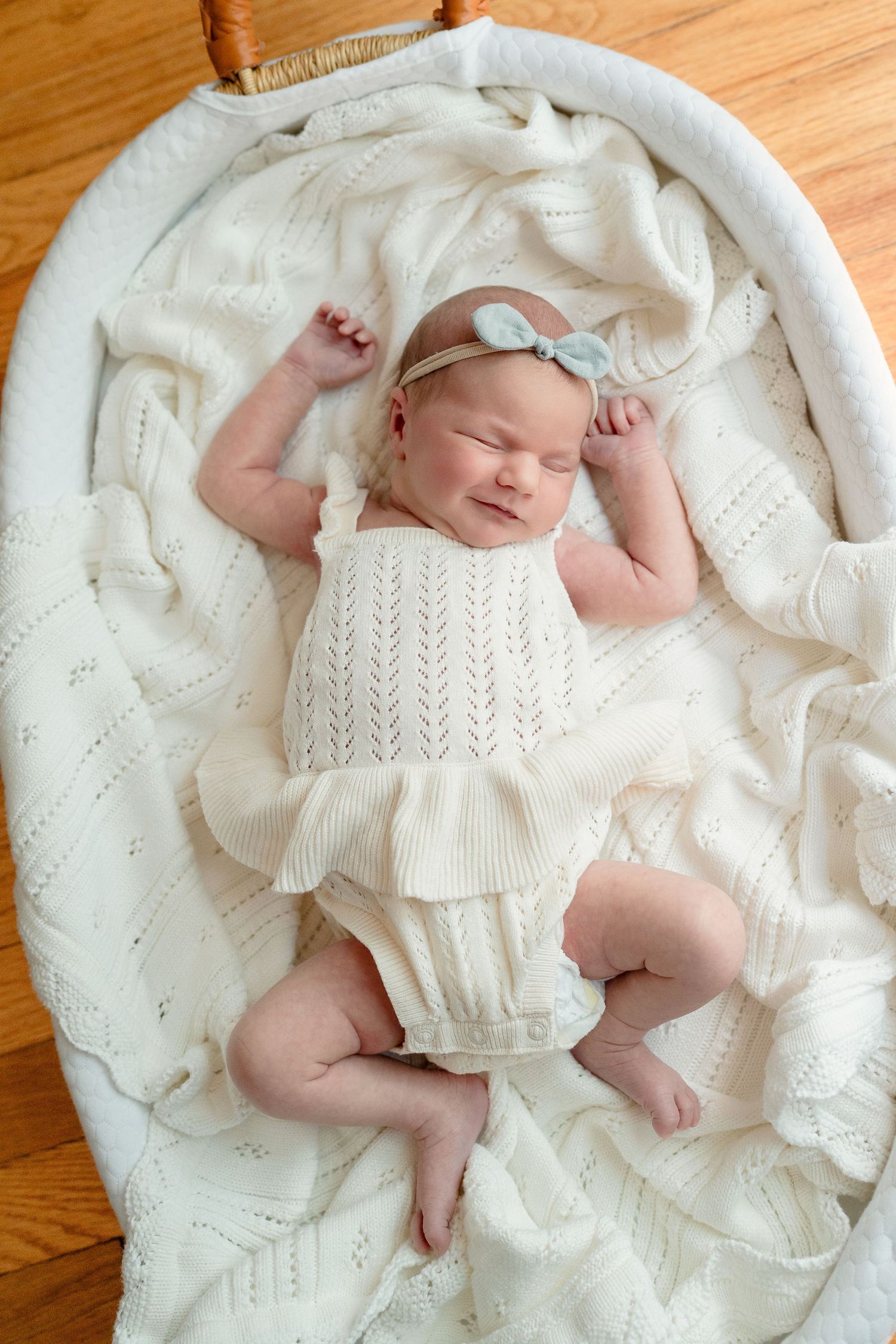 Newborn baby girl in white knit romper and bow headband sleeping peacefully in a white wicker bassinet.
