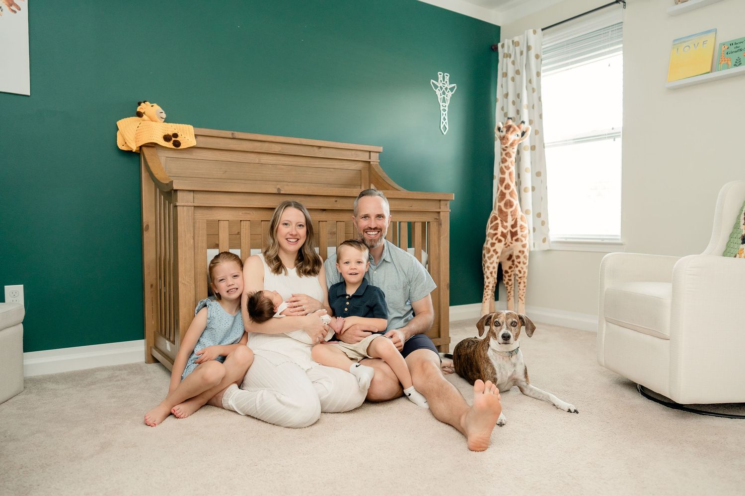 Happy family of five with newborn baby poses in front of green nursery wall with wooden crib and giraffe decor in Metro Detroit, MI