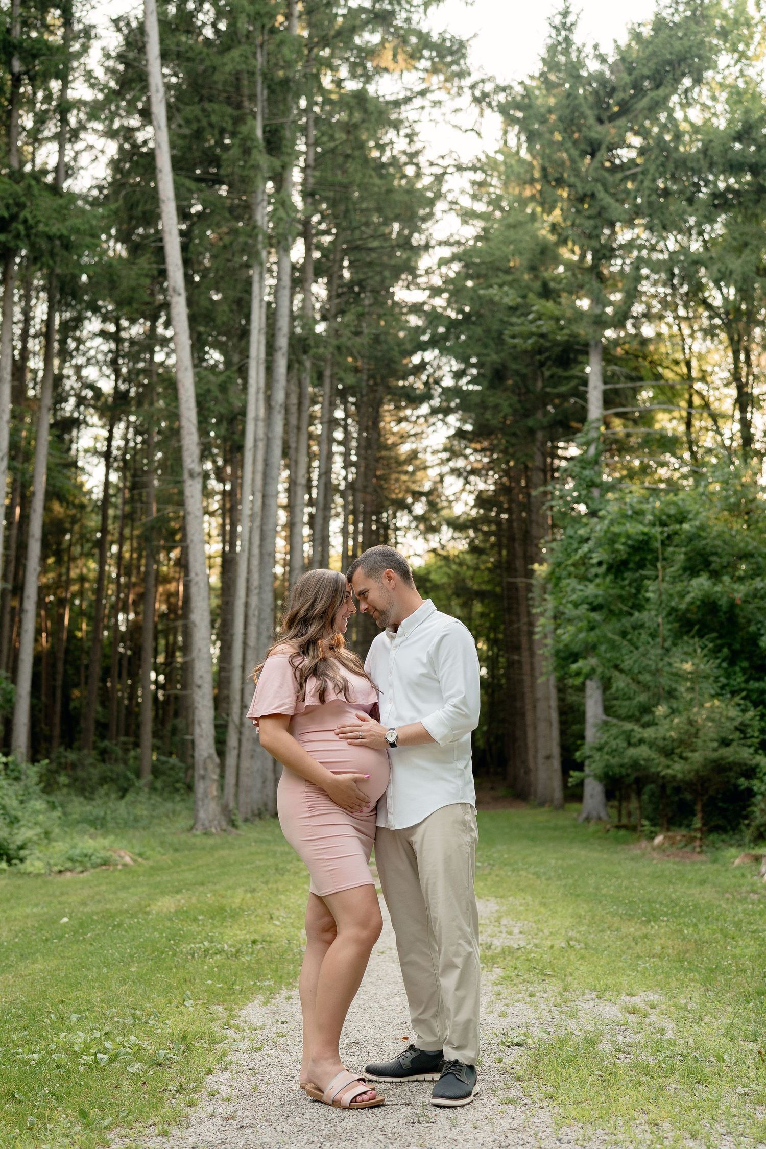 Expecting couple embracing on a gravel path surrounded by tall pine trees during golden hour maternity shoot.