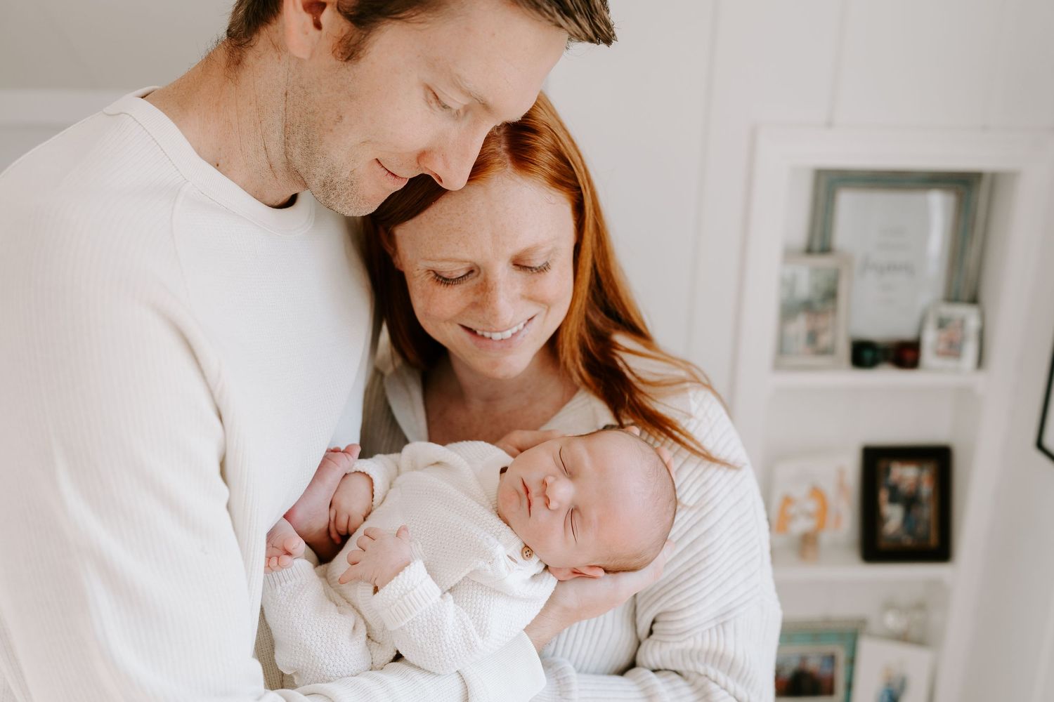 Happy couple in white clothing cradling their sleeping newborn baby together.