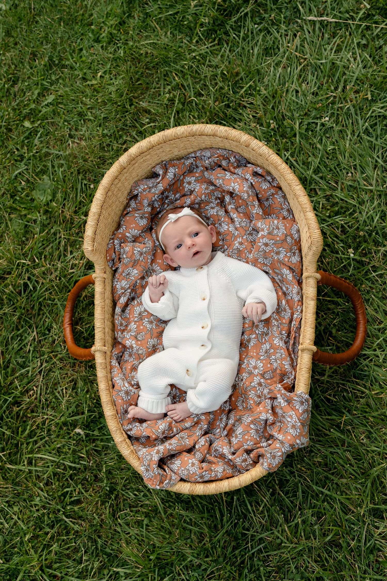 Newborn baby in white outfit lying in a wicker Moses basket on green grass with floral blanket.