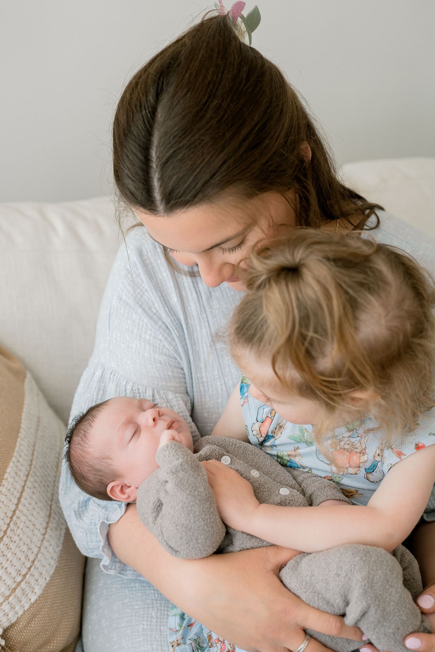 Loving mother sitting on sofa holding sleeping newborn while toddler sibling gazes adoringly.