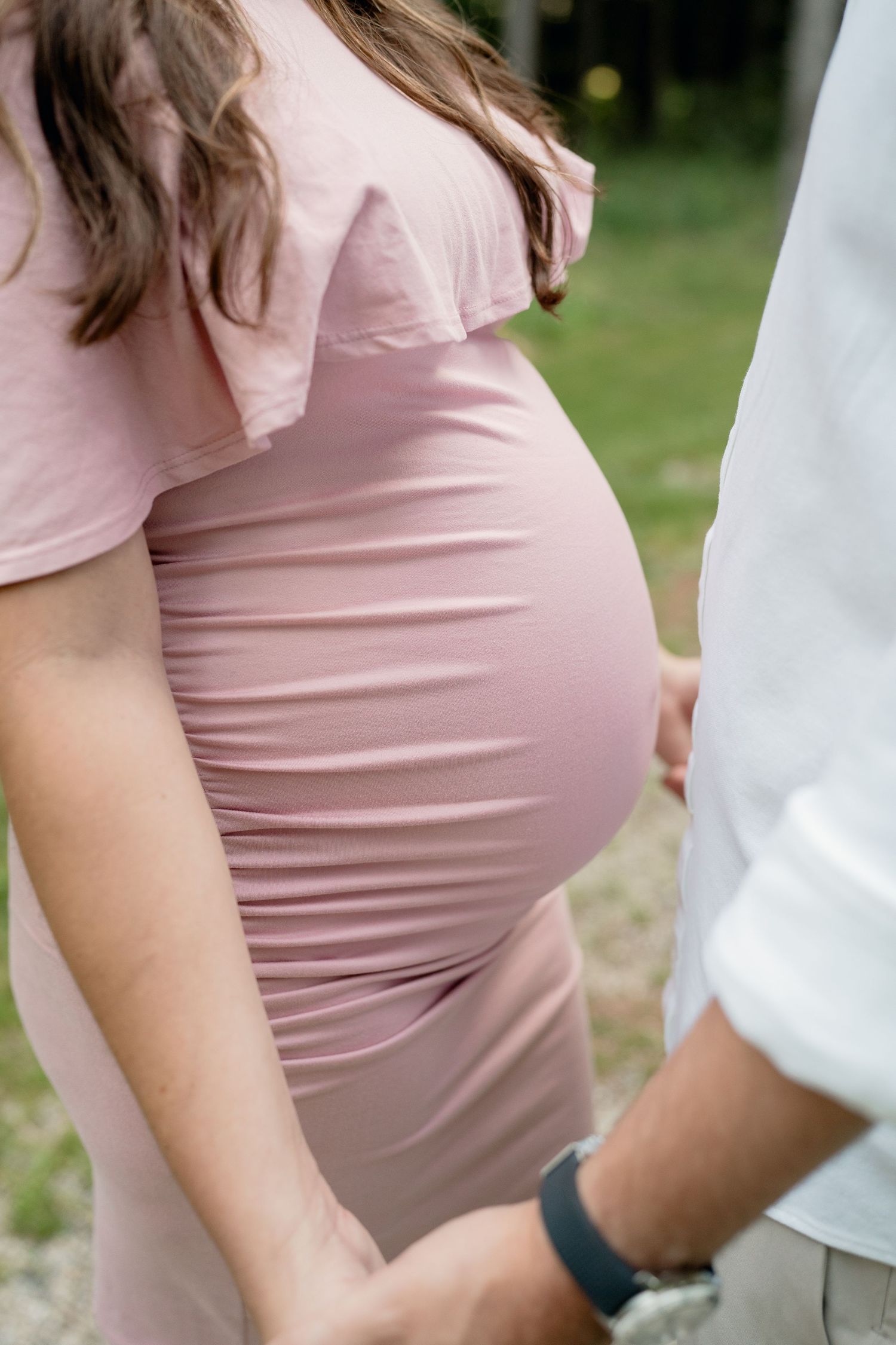 Close-up of a pregnant womans baby bump in a blush pink fitted maternity dress holding partners hand outdoors.