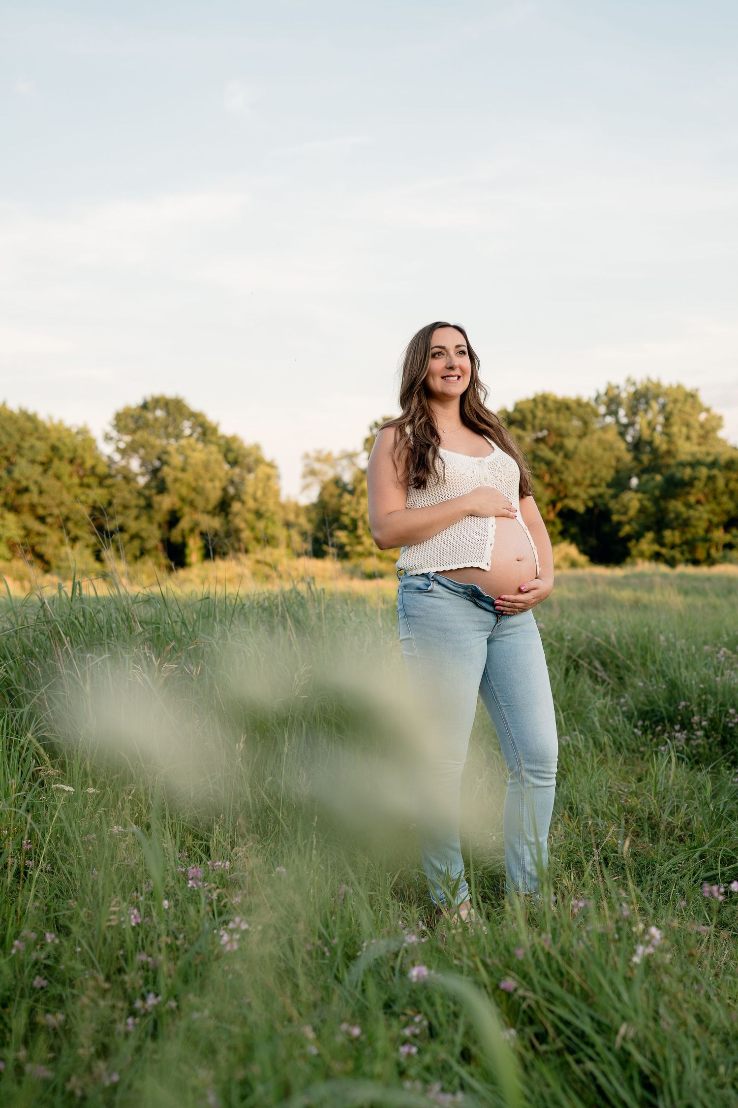 Smiling pregnant woman in white top and jeans cradles bump in green field.