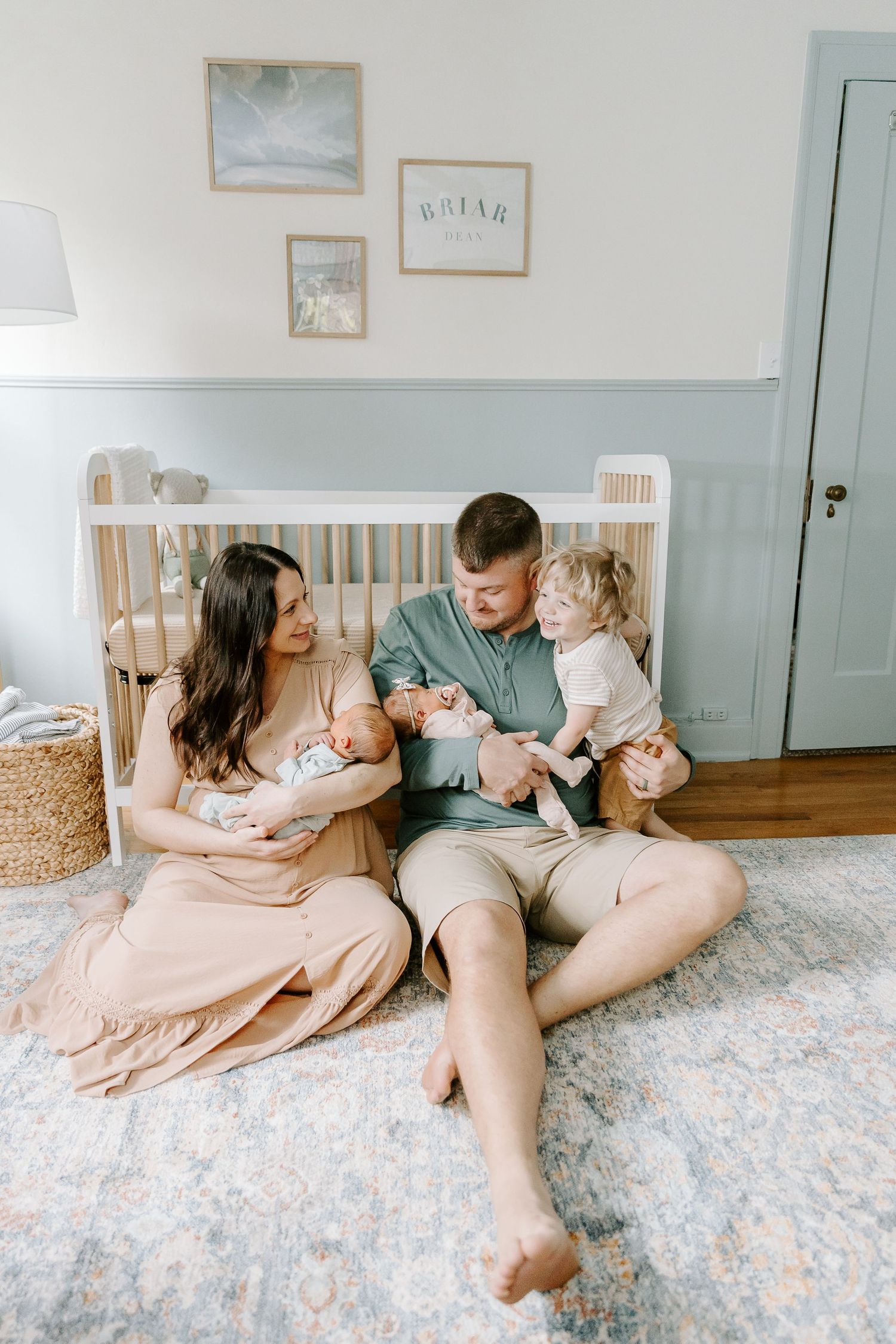 Young family with toddler and twin newborns sitting on nursery floor in front of white crib.