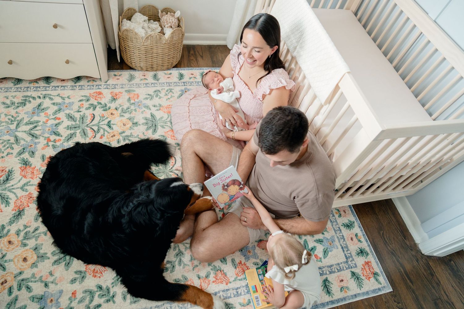 Young family with newborn baby and Bernese Mountain Dog sitting on floral rug in nursery reading a book.