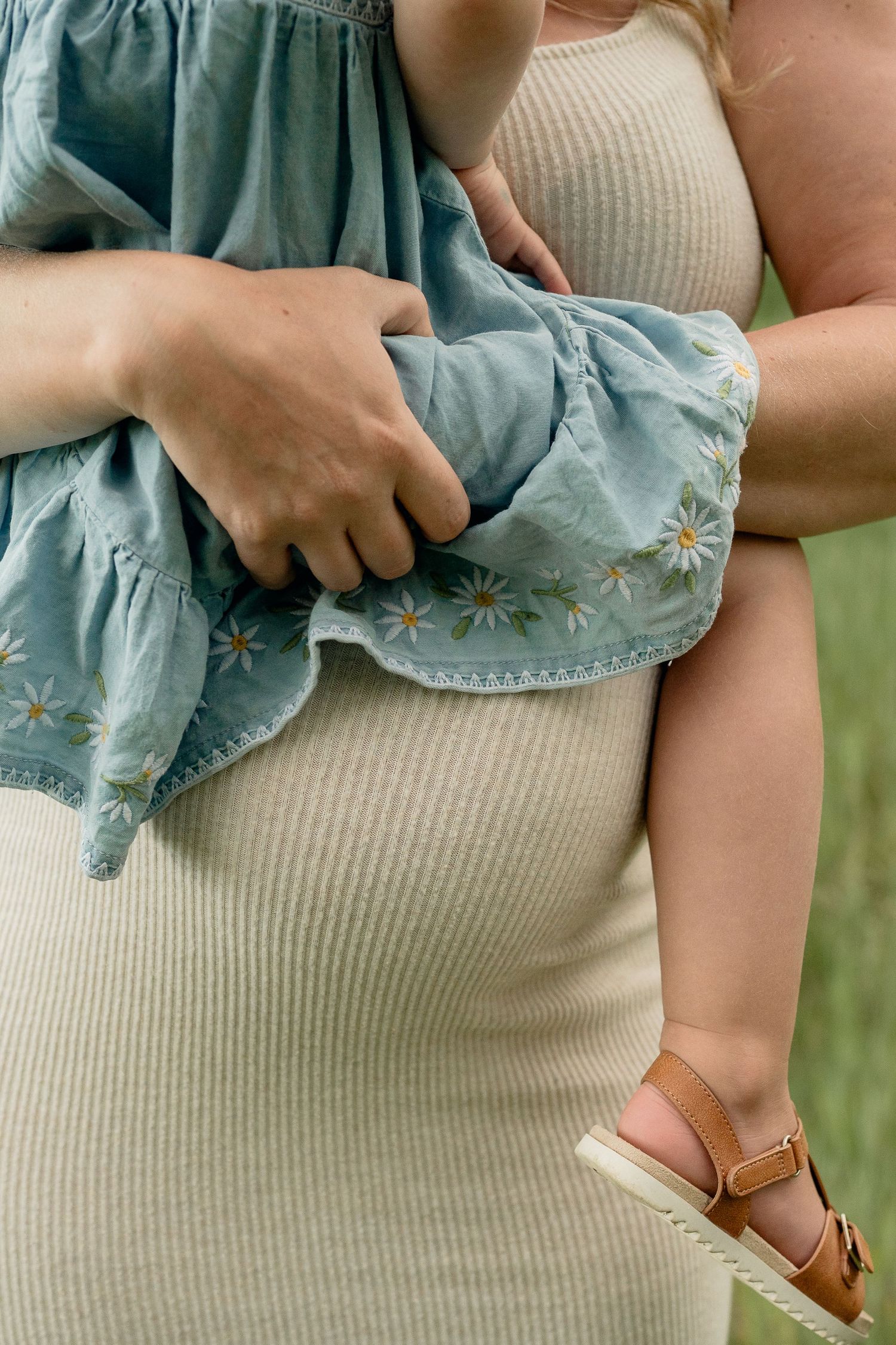 Pregnant woman in ribbed beige dress holding a toddler in a blue daisy embroidered dress outdoors.