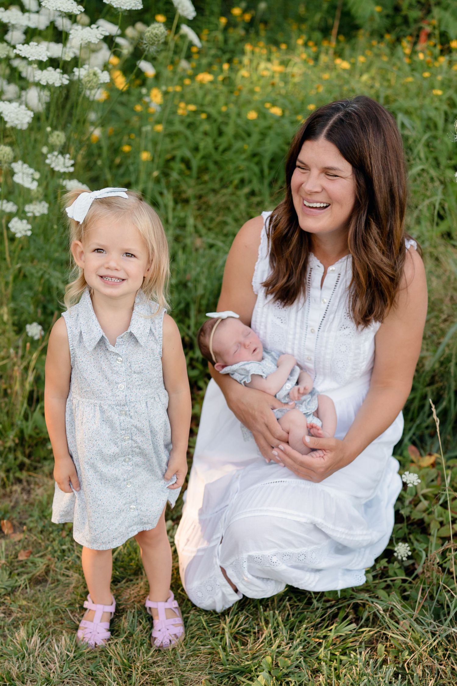 A laughing brunette woman in white dress holds newborn baby beside smiling blonde toddler girl in wildflower field.