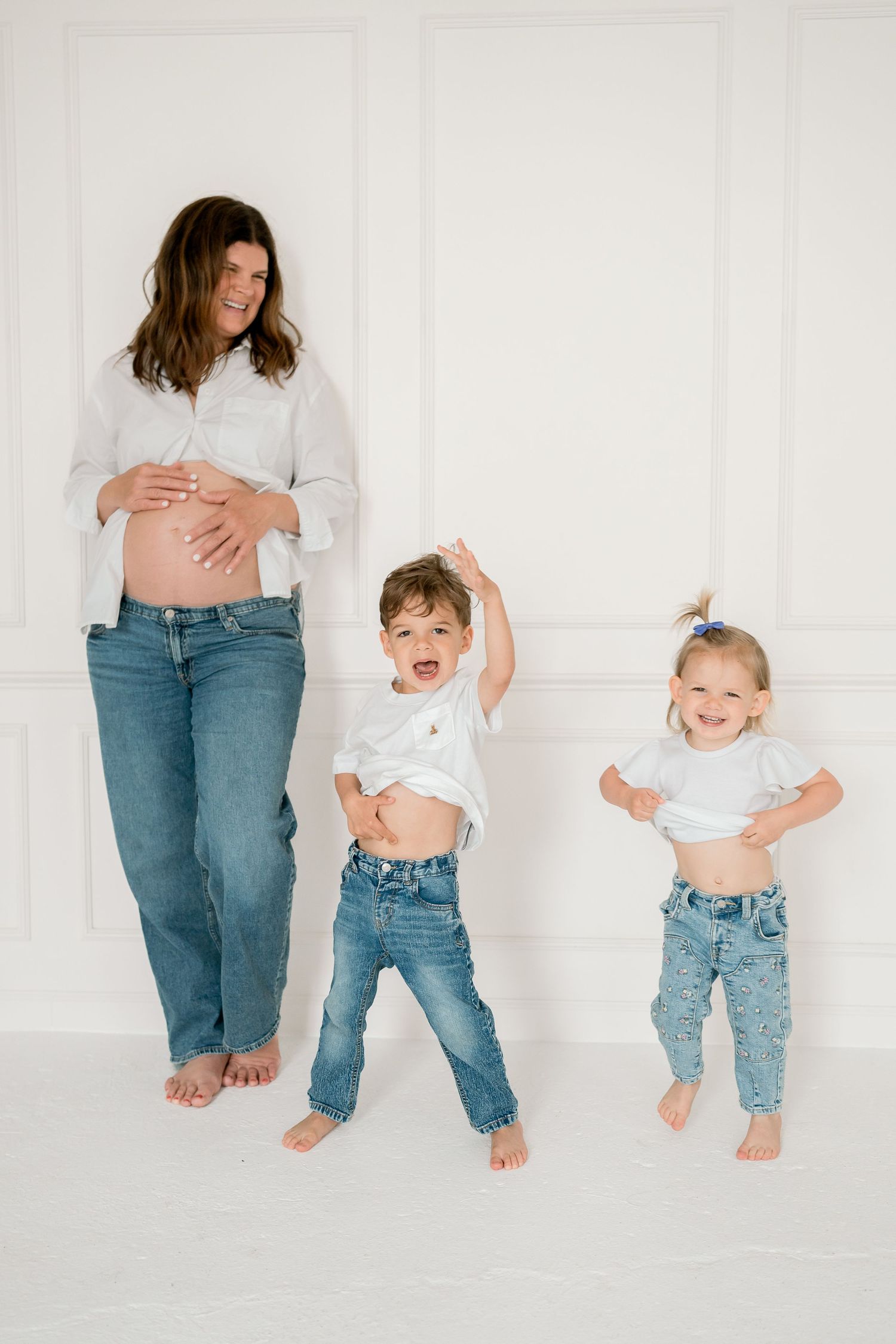Pregnant woman in white shirt and jeans smiling as two young children playfully lift their shirts beside her.
