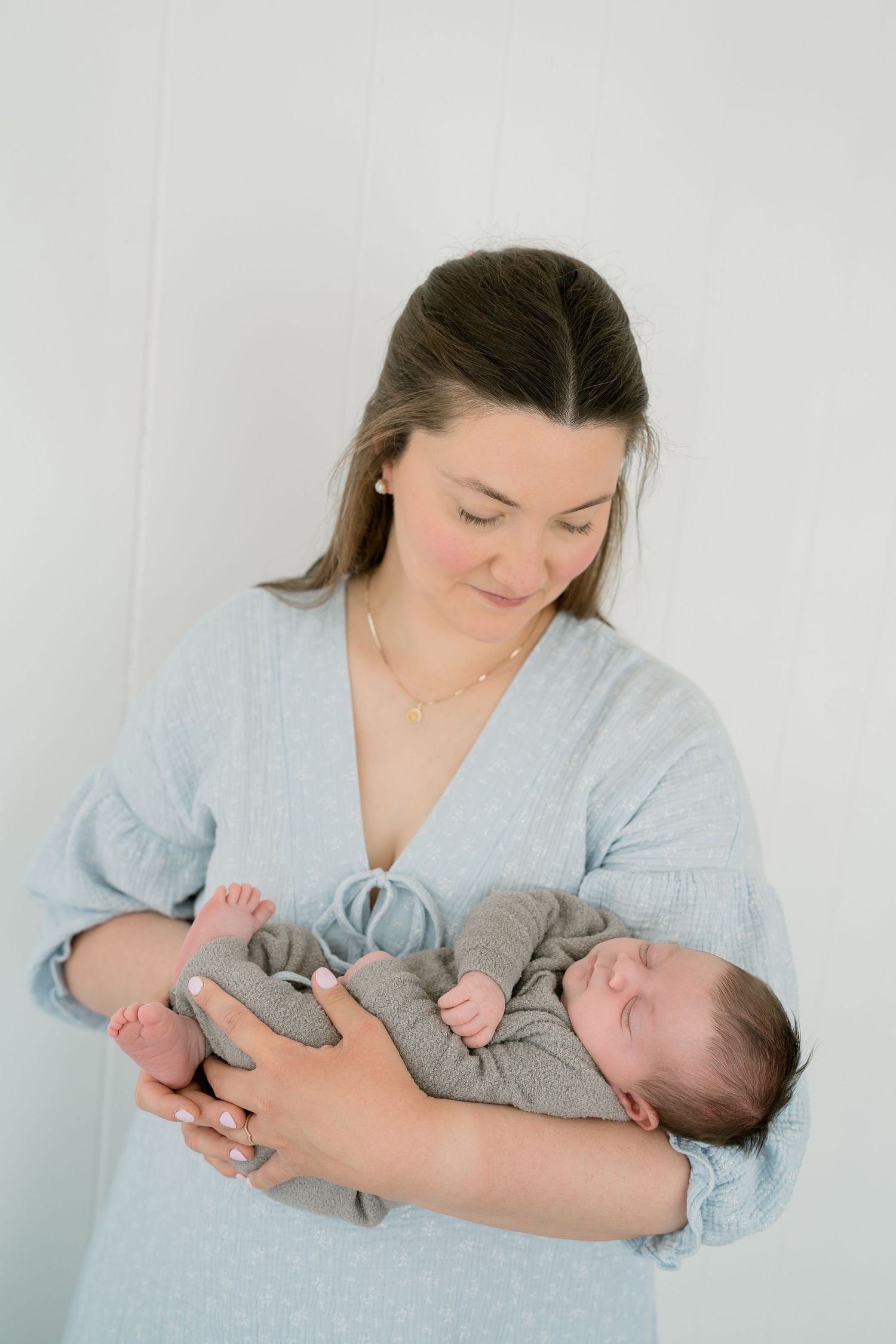 A mother in a light blue dress tenderly holds her sleeping newborn baby against a soft white background.