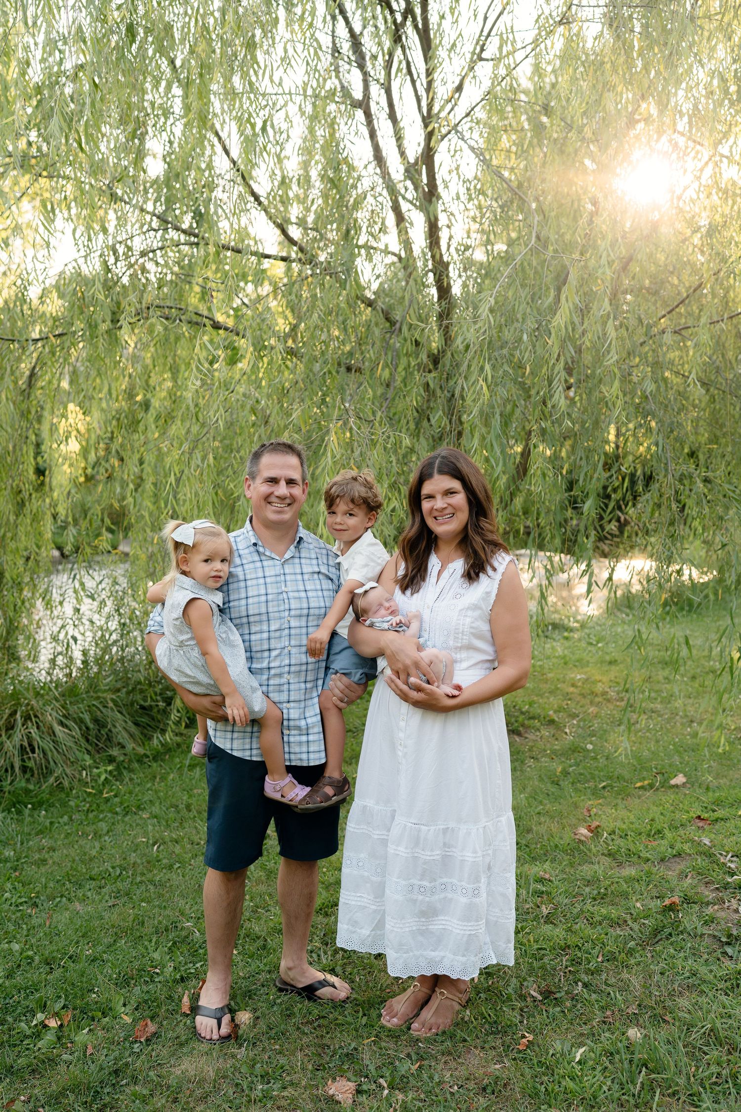 Happy family of four posing outdoors under a weeping willow tree by a pond during golden hour.