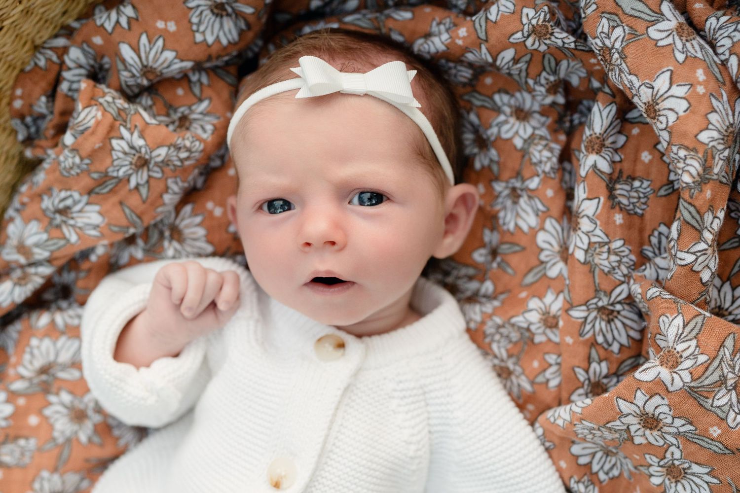 A newborn baby with blue eyes wearing a white knit cardigan and bow headband, lying on a floral rust-colored blanket.