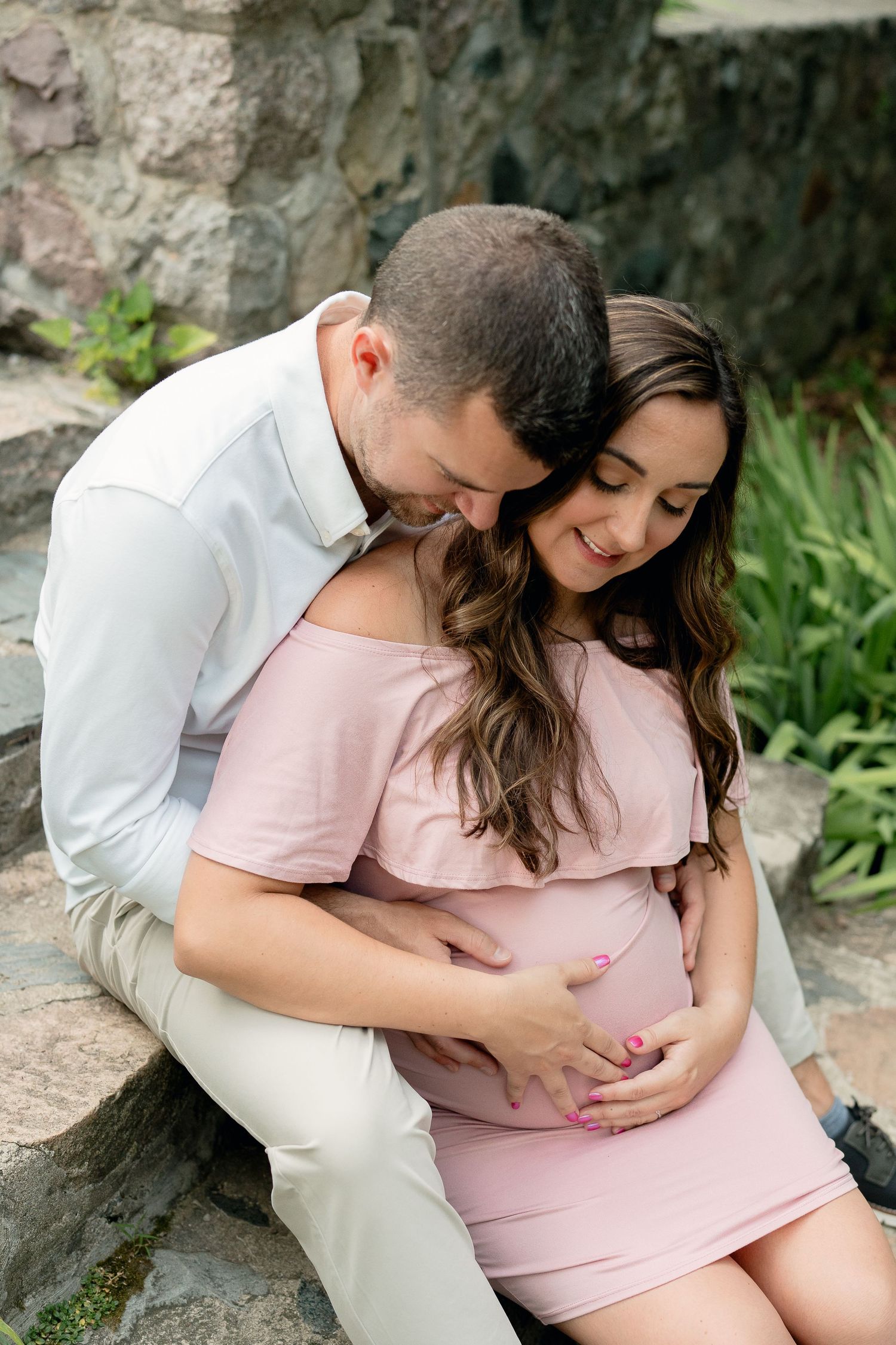 Couple tenderly embracing on stone steps outdoors, mans hands on pregnant partners baby bump during maternity shoot.