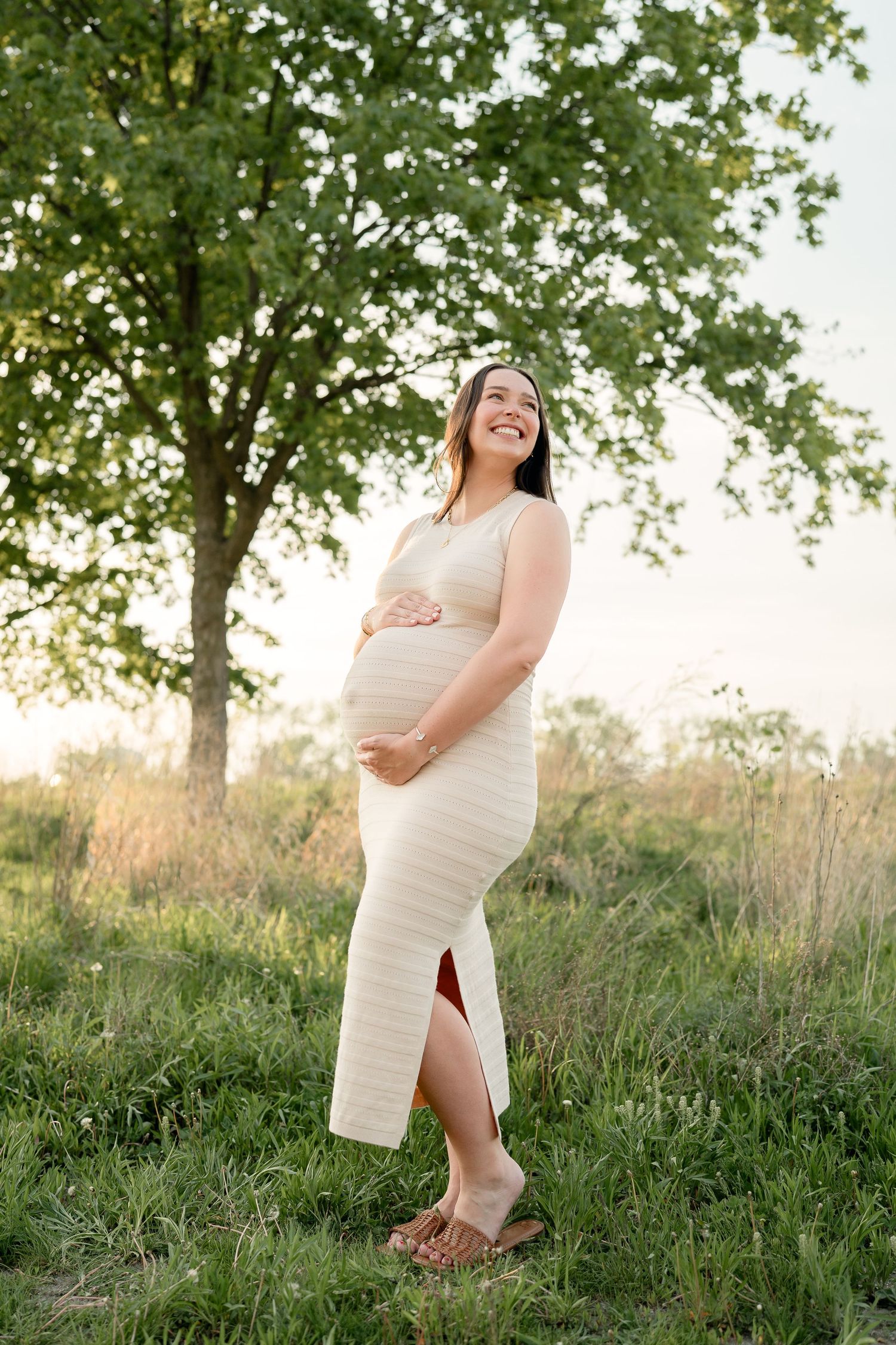 Glowing pregnant woman in cream ribbed dress cradling belly near tree at golden hour.