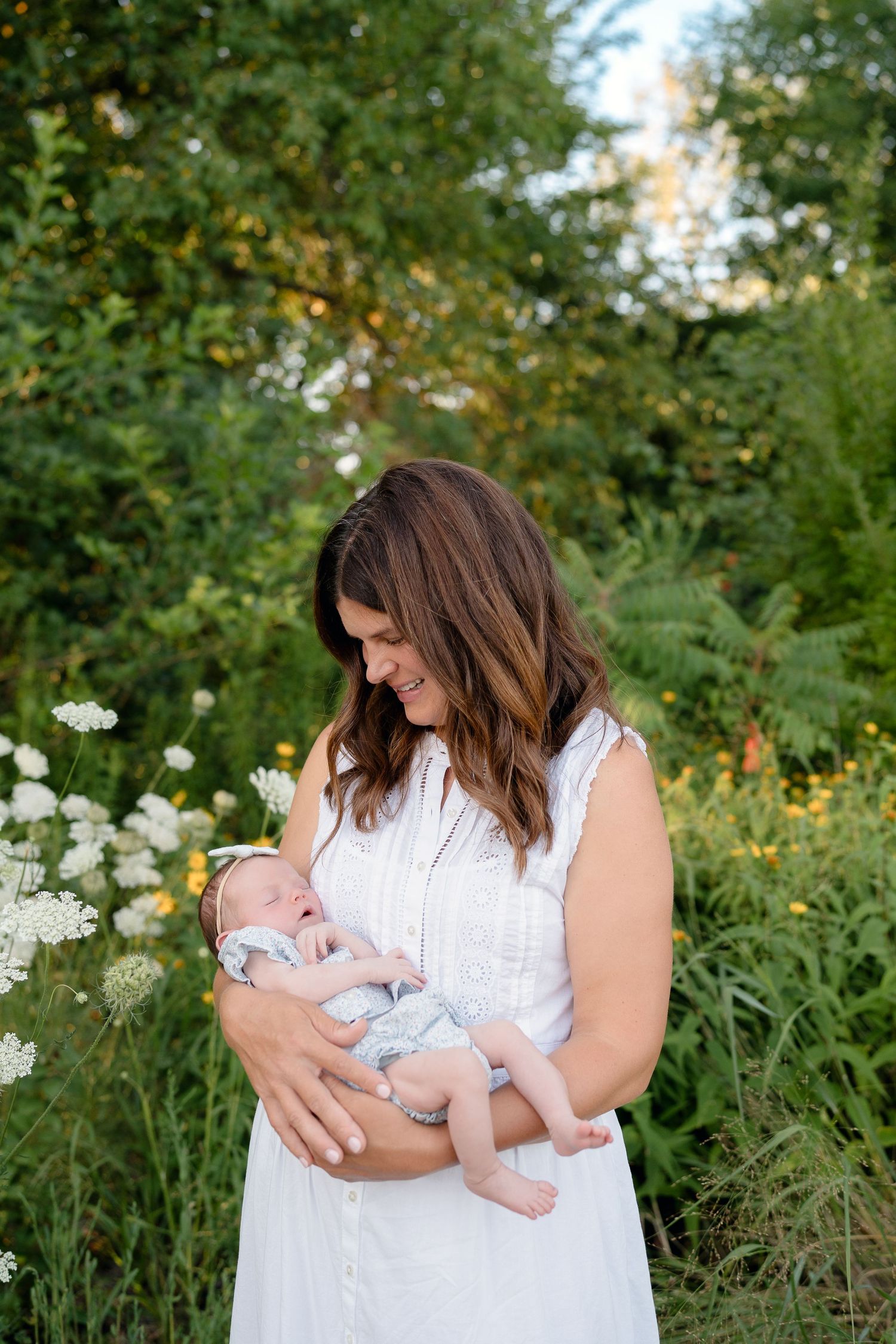 A smiling mother in a white dress cradles her newborn baby outdoors among wildflowers and lush greenery.