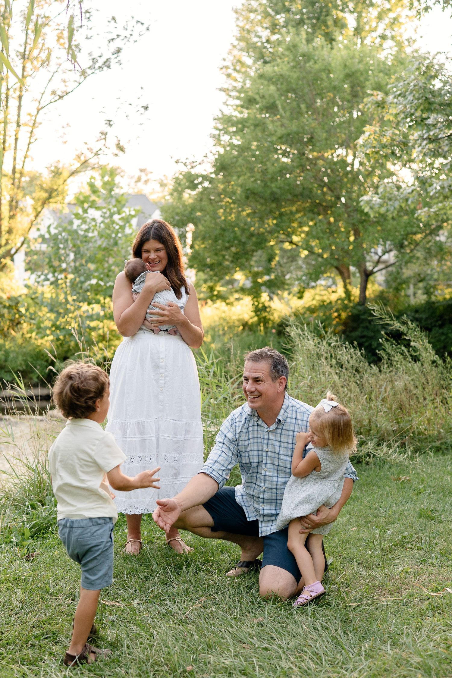 Happy family with father, mother holding newborn, toddler son, and young daughter playing together in sunny park.