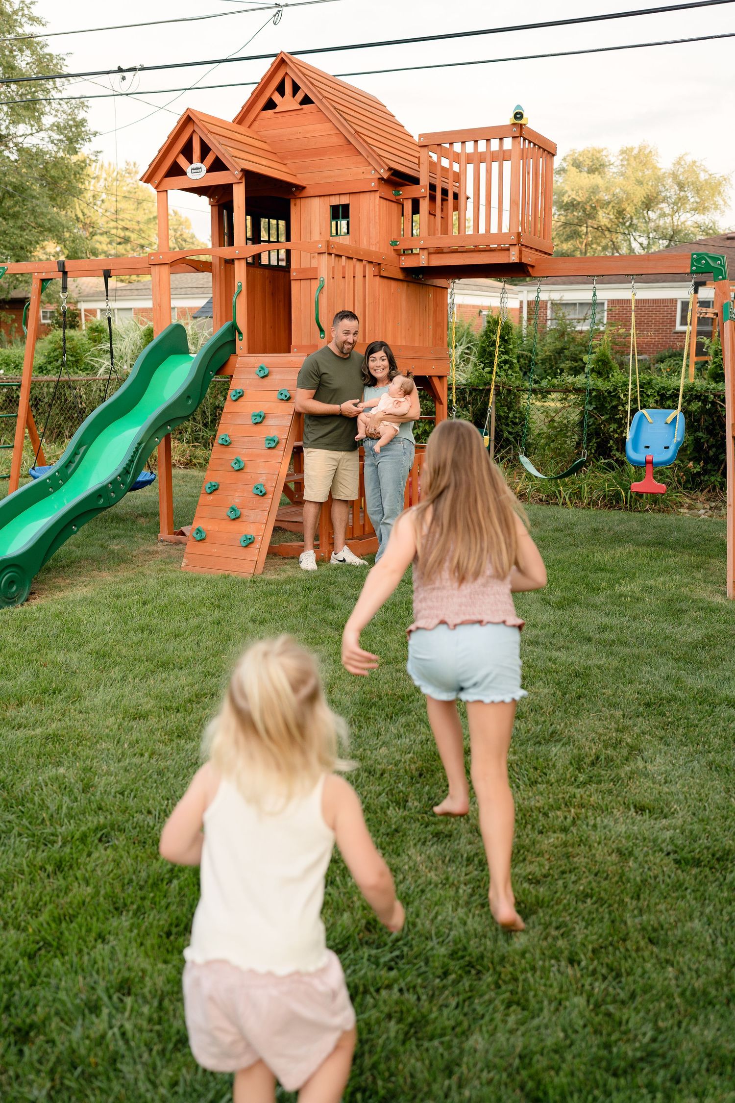 Family enjoying backyard playset with slide, climbing wall, and swing set on a green lawn.
