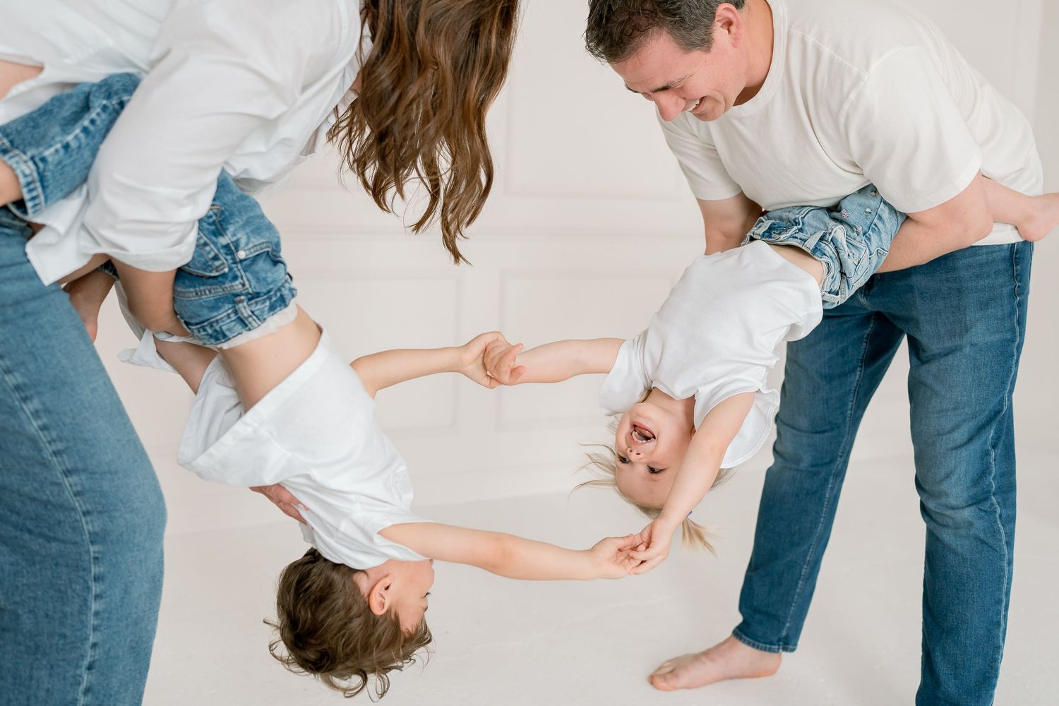 Playful family photo with parents swinging laughing children upside down in bright white studio.